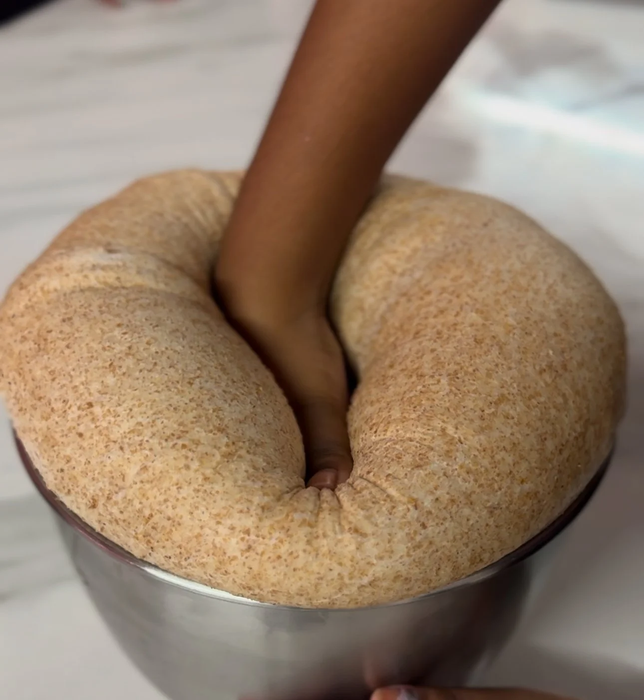 A hand is pushing down on a large, round, light brown bread with a soft, speckled surface, placed in a metal bowl.