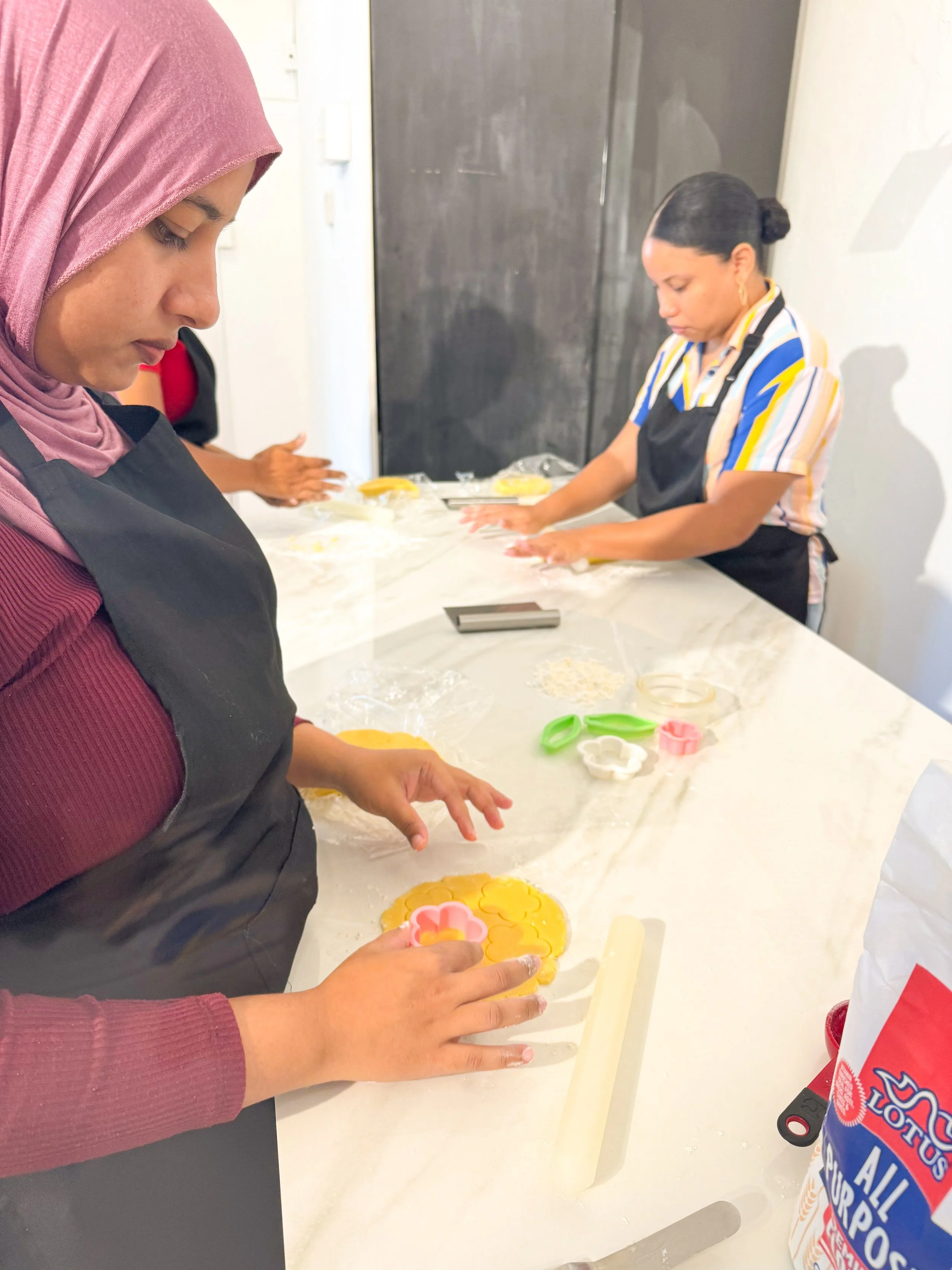 Two women wearing aprons are decorating cookies with cookie cutters and icing on a white kitchen counter.