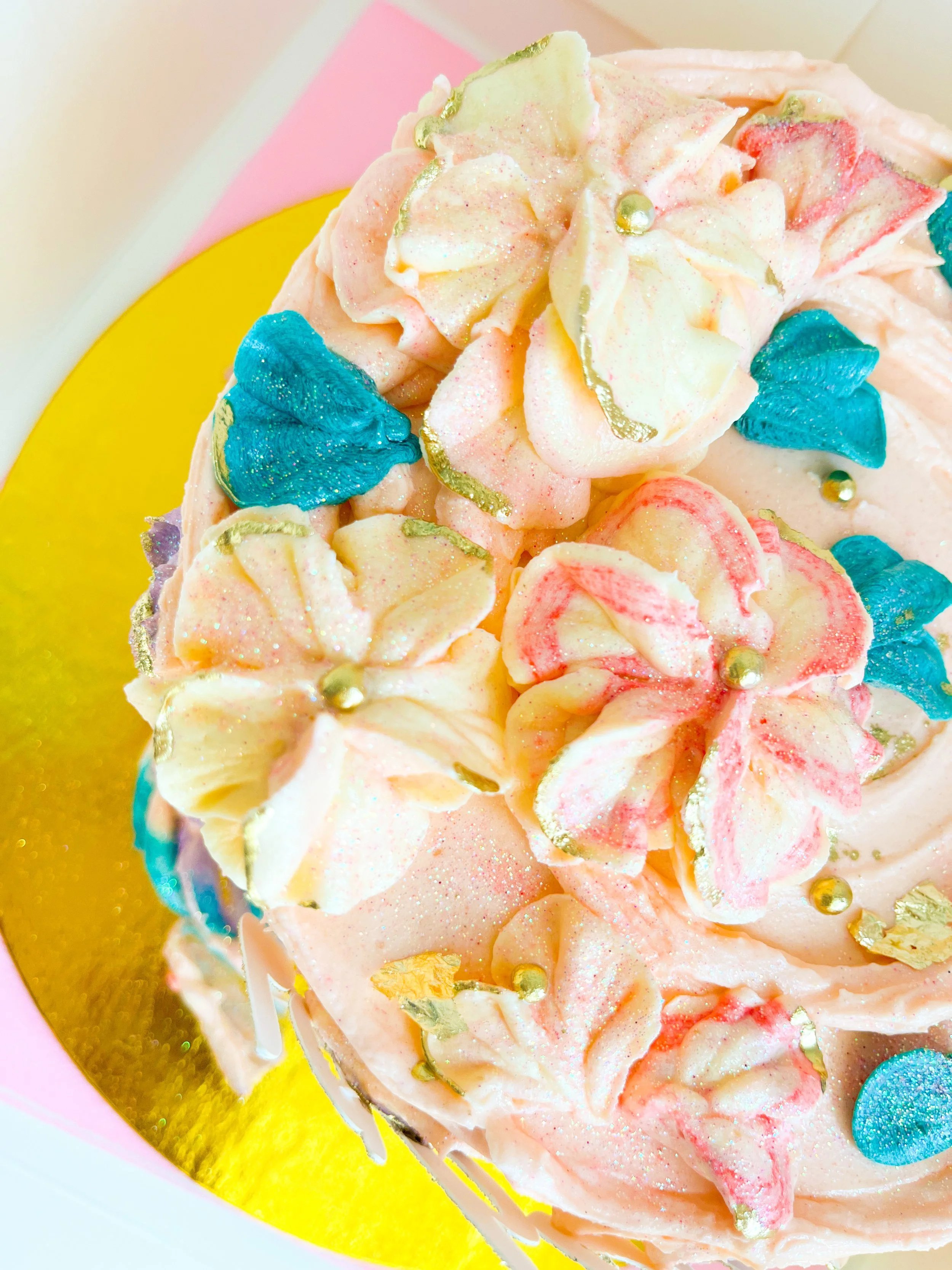 Close-up of a decorated cake with pink buttercream, blue and gold decorative accents, and edible flowers on top, placed on a yellow and pink plate.