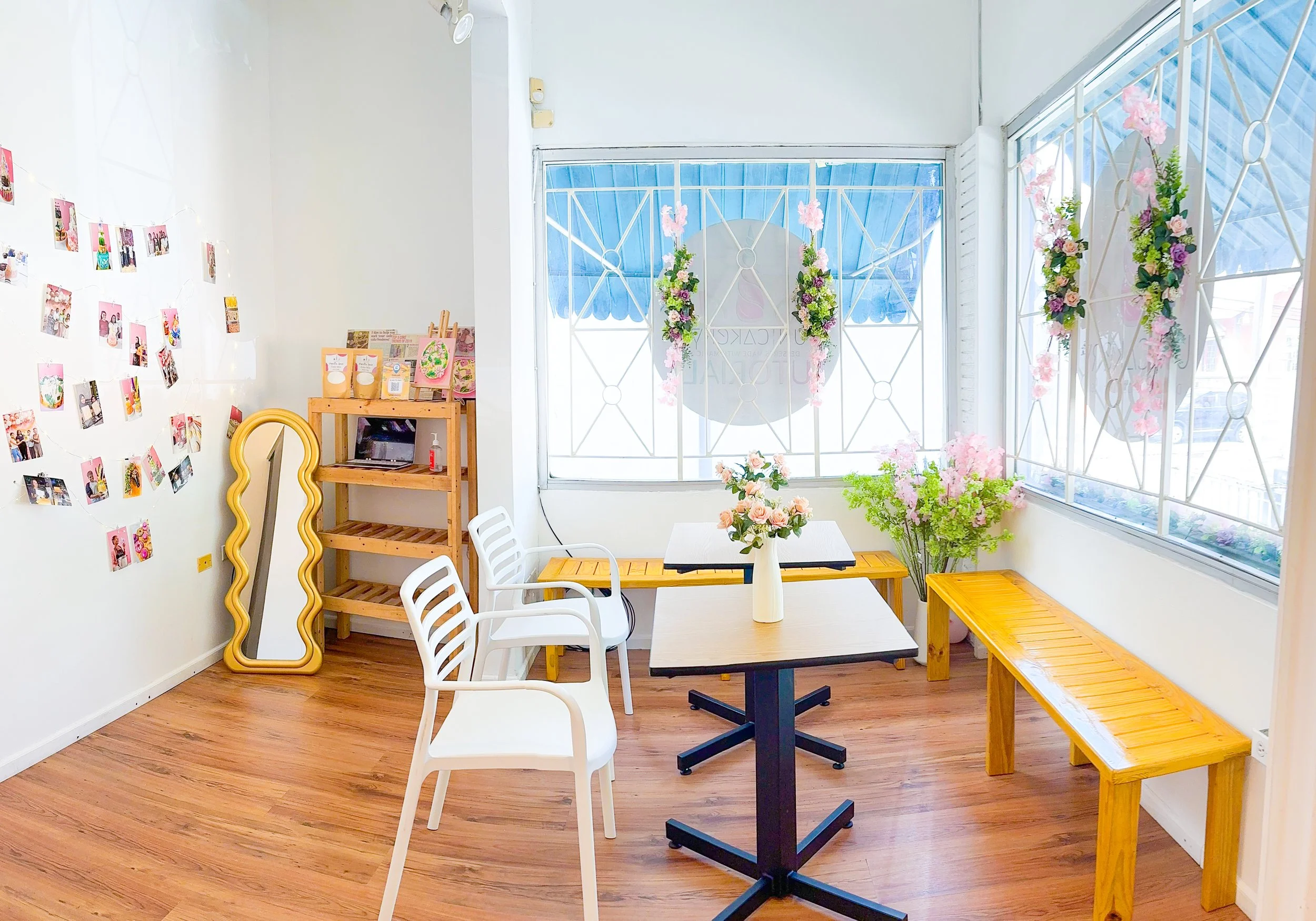 A cozy cafe corner decorated with pink and purple flowers, a small white table with white chairs, a wooden shelf with craft supplies, and a wall with photo clips. Large windows with white lattice bars are adorned with floral arrangements, letting in natural light.