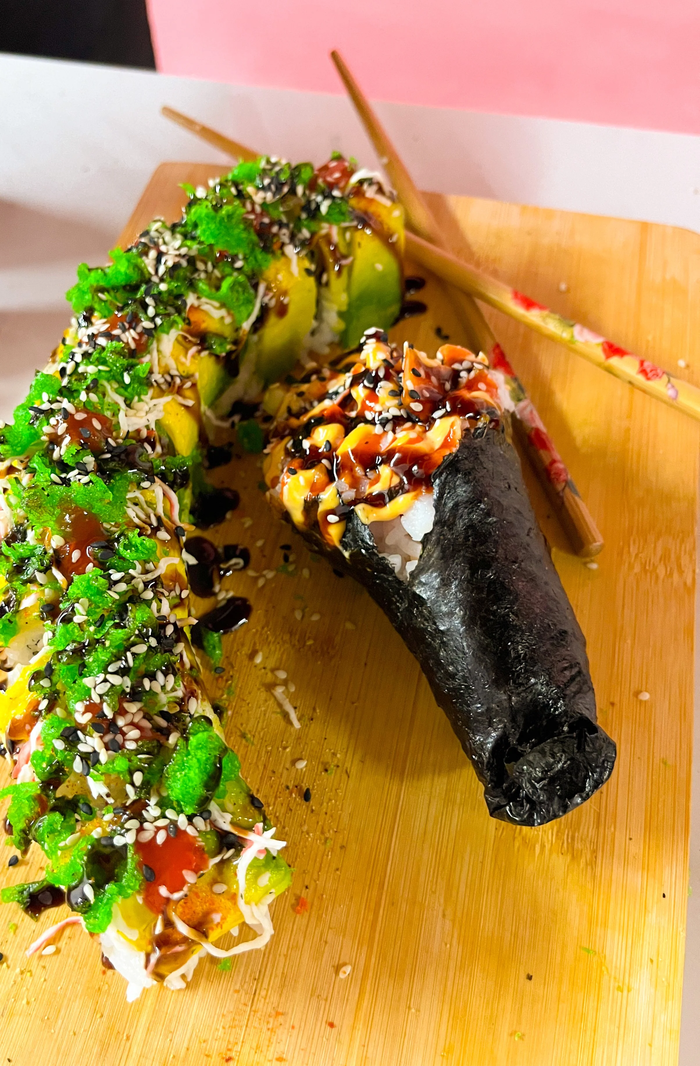 Close-up of sushi rolls and a sushi hand roll on a wooden board, garnished with sesame seeds, sauces, and colorful toppings.