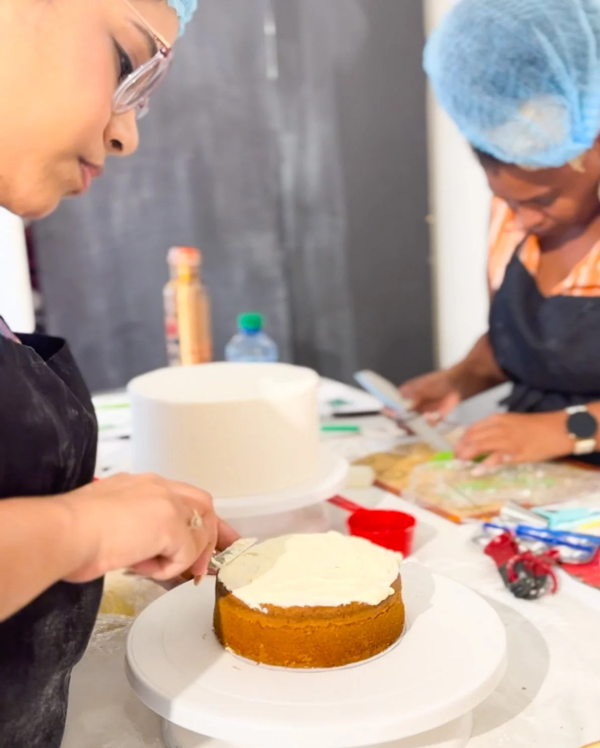 Two women decorating a cake in a kitchen or bakery setting, with various baking tools and ingredients on the table.
