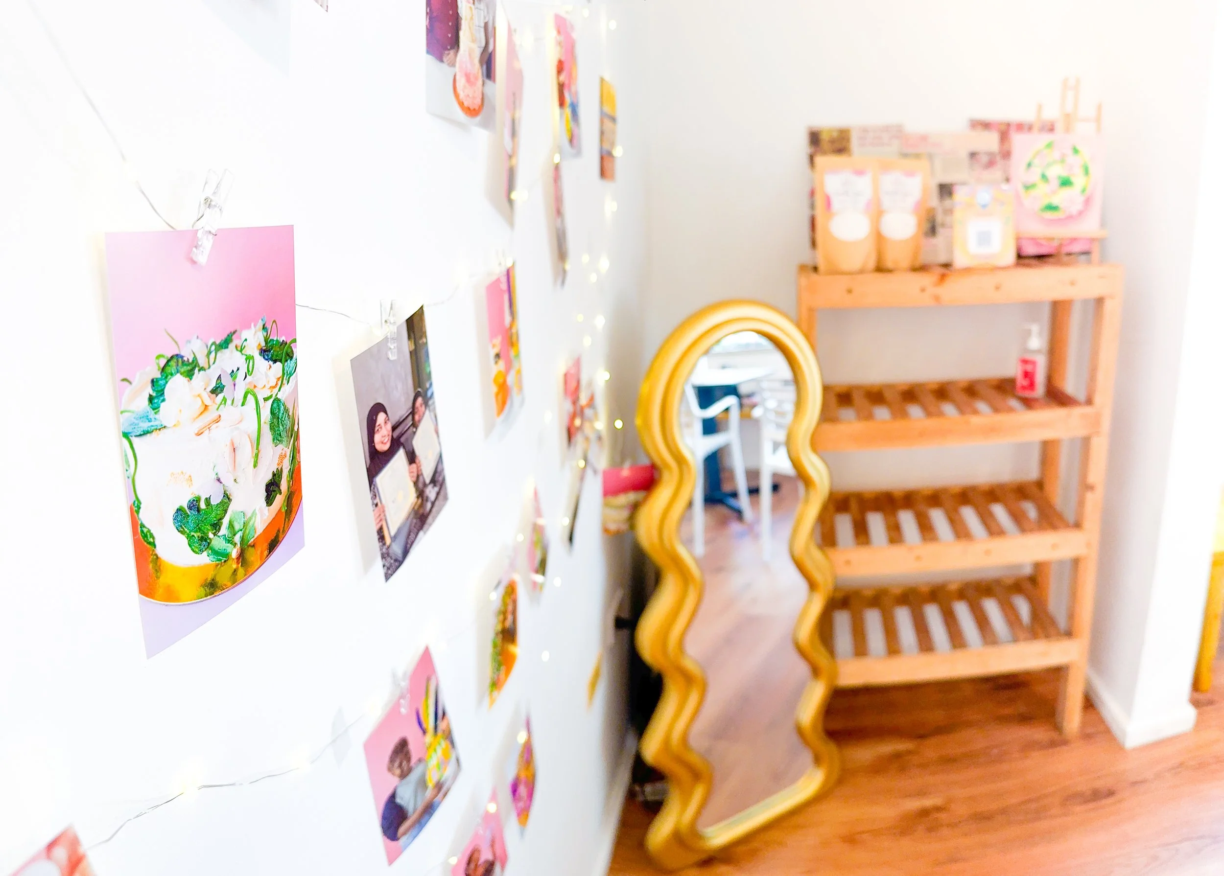 A wall decorated with various colorful pictures, fairy lights, and pink paper notes, next to a wooden shelf with boxes and a yellow heart-shaped mirror.