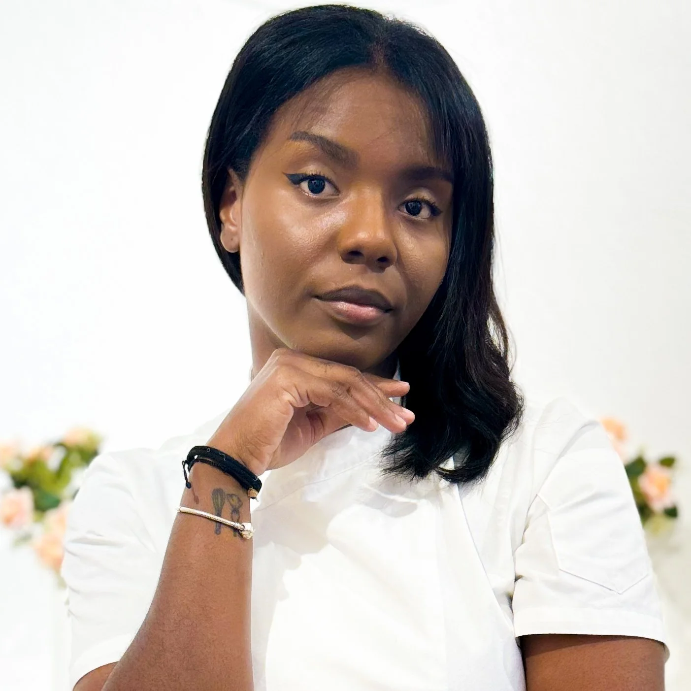 A woman with wavy black hair, wearing a white shirt, resting her chin on her hand, with bracelets on her wrist, standing in front of a plain white background with some blurred flowers.