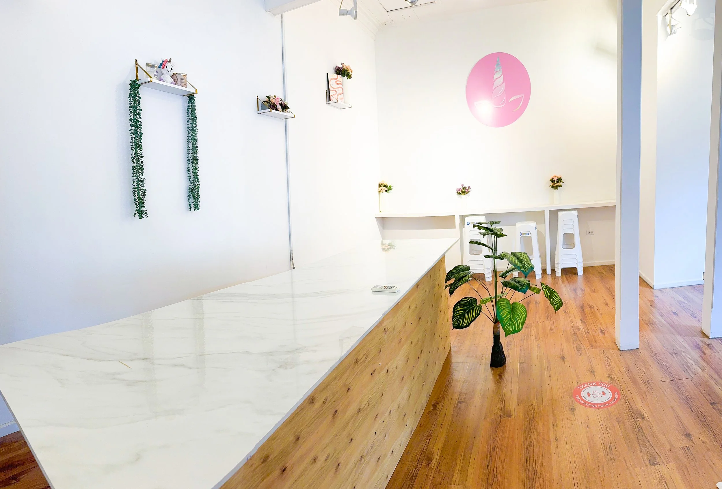 Interior of a bright, modern room with white walls, a wooden floor, and minimal decorations including small flower arrangements and hanging shelves with a plush unicorn toy and plants. There is a pink circular logo with a unicorn horn on the wall. A small potted plant is on the floor, and a social distancing floor sticker reads "Thank you for practicing social distance."