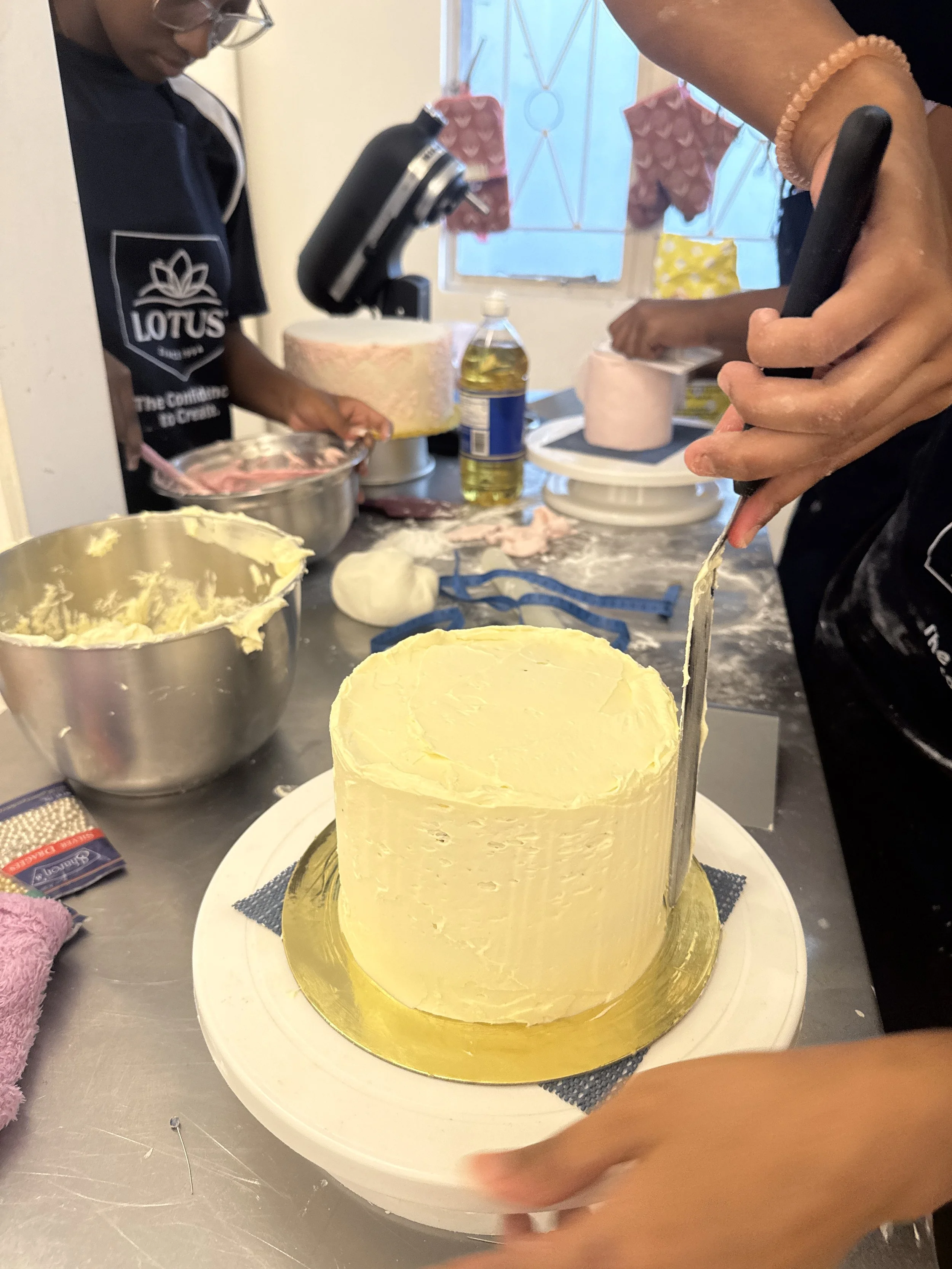 A person is cutting a yellow cake with a large knife, with another person in the background preparing ingredients, in a kitchen.