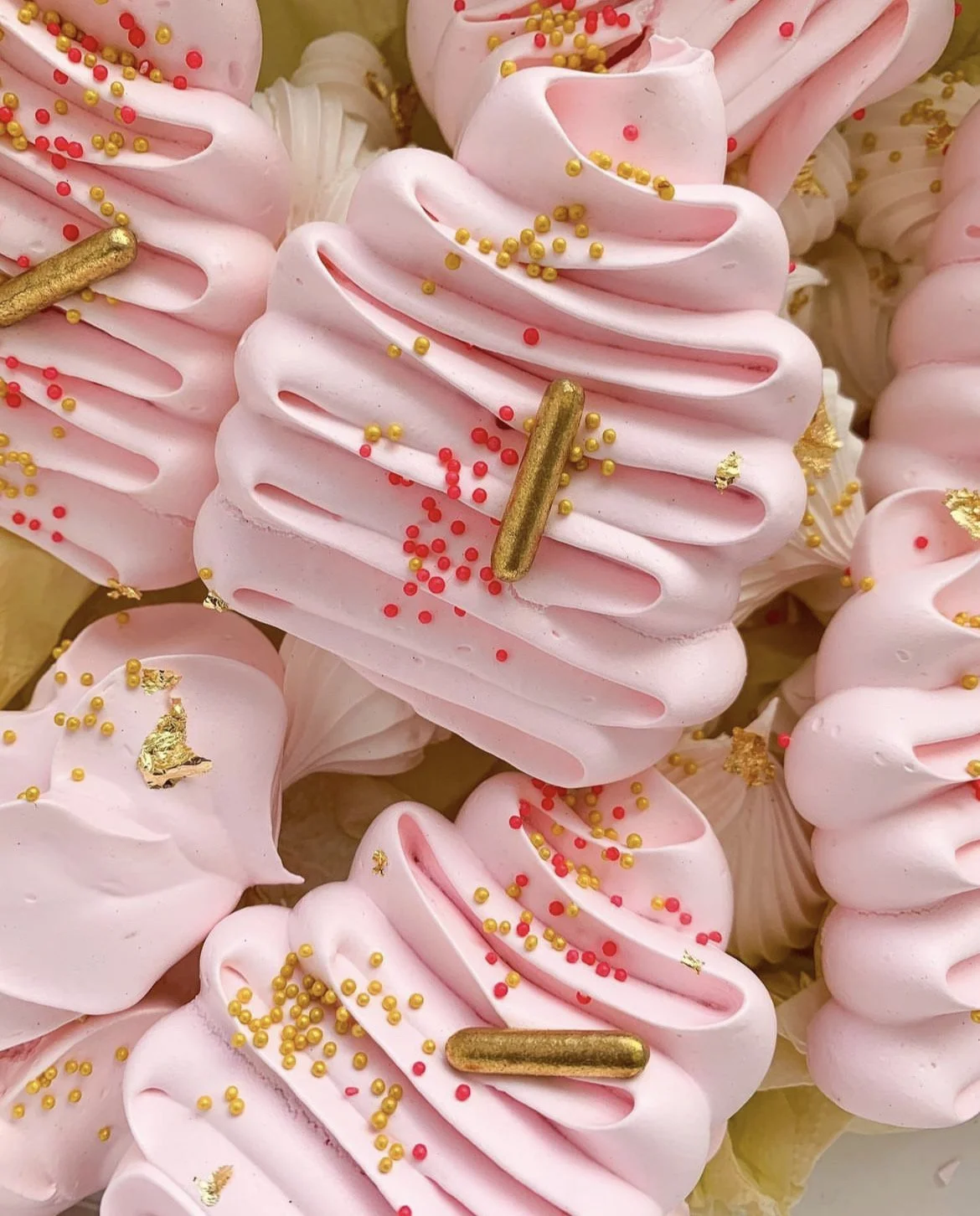 Pink meringue cookies decorated with red and gold sprinkles, gold leaf accents, and gold and pink elongated sprinkles.