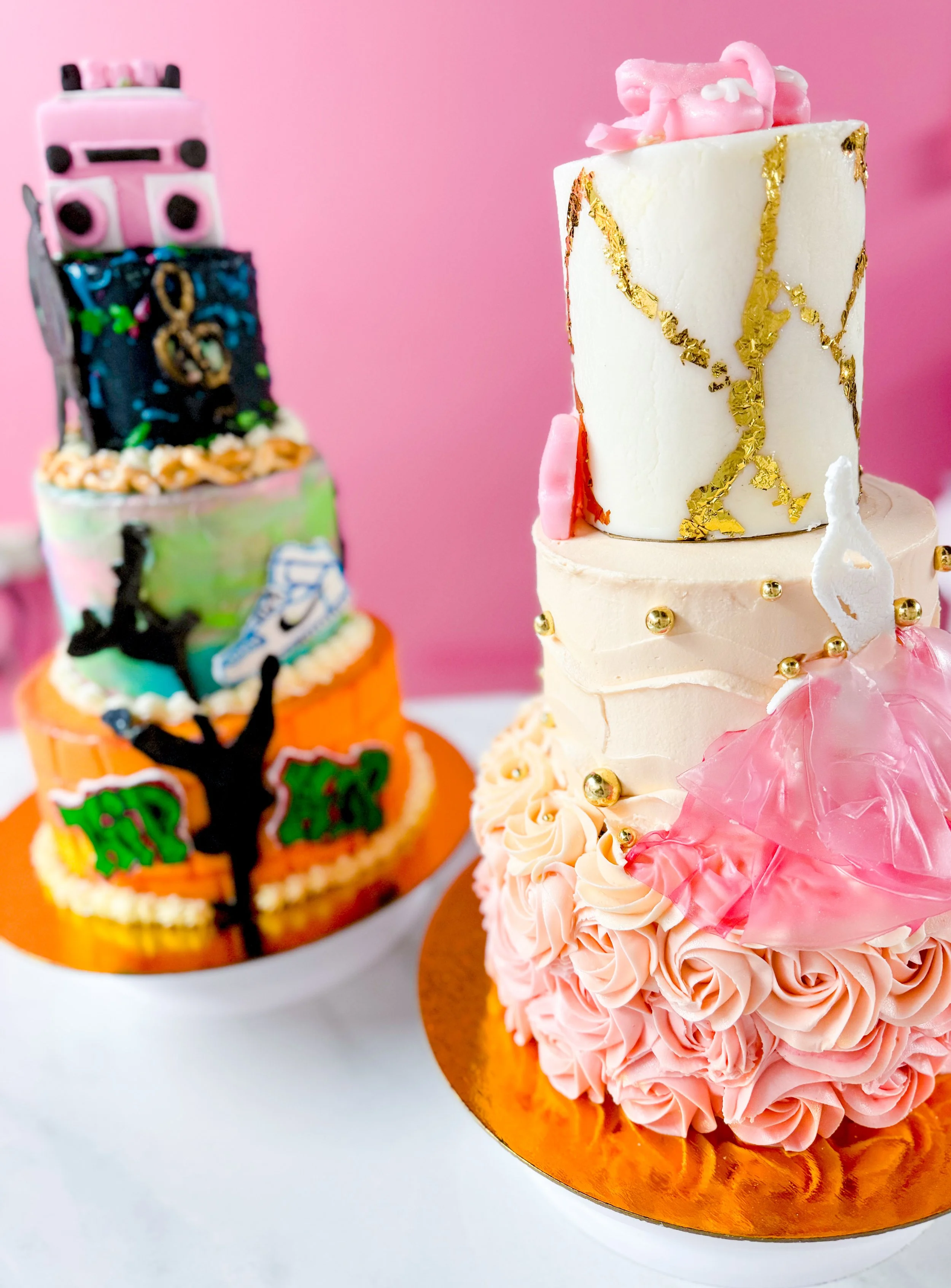 Close-up of a pink birthday cake decorated with pink roses and gold leaf, with a smaller cake in the background decorated similarly.