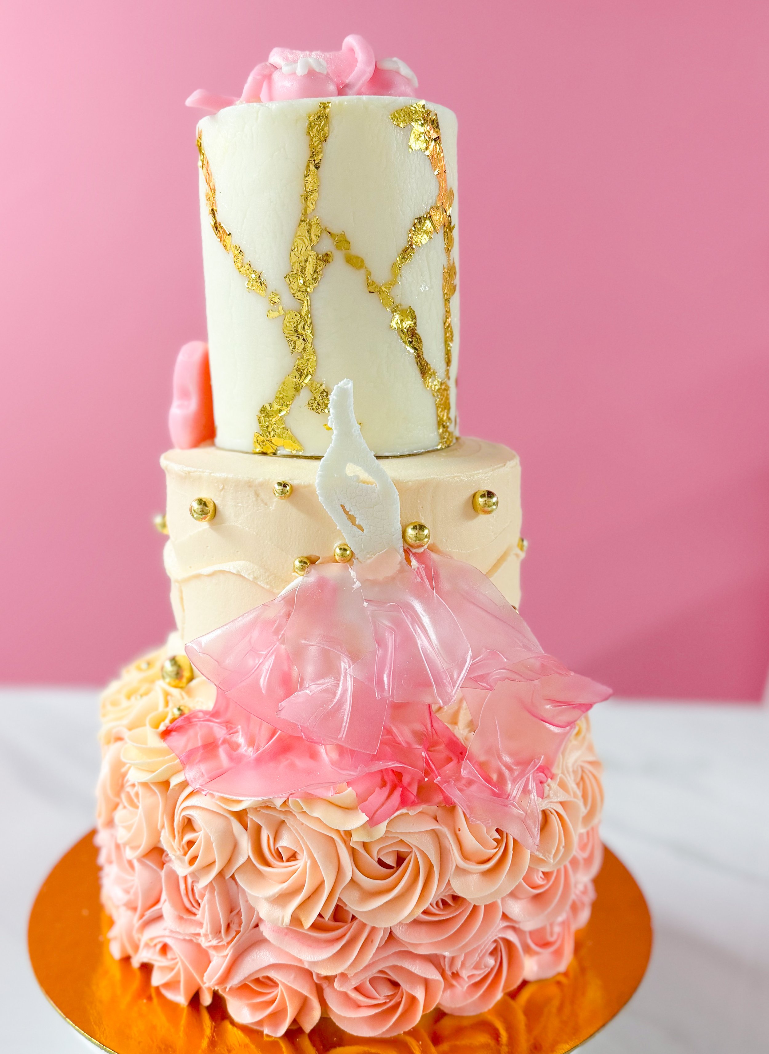 A tiered cake decorated with pink and white frosting, gold leaf accents, and pink chocolate decorations on top. The cake has pink ruffled frosting at the base and a light pink backdrop.