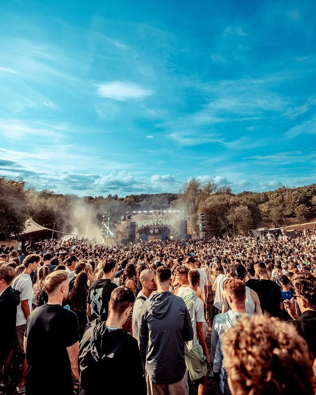 Crowd of people attending an outdoor music festival with a stage in the background, trees, and a blue sky.