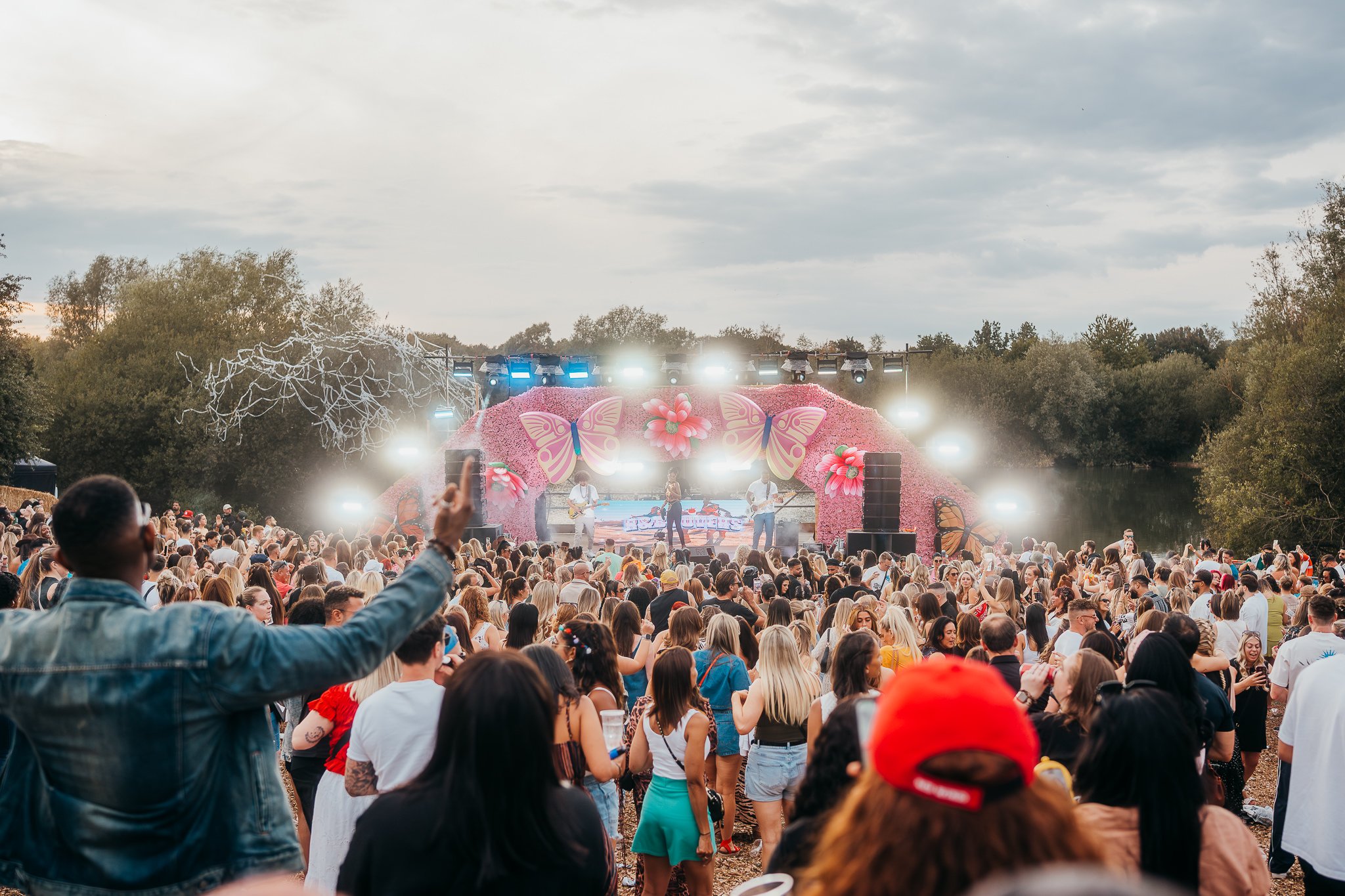 Large outdoor concert with a stage decorated with pink flowers and butterflies, an audience gathered nearby, some taking photos, overlooking a river and surrounded by trees.