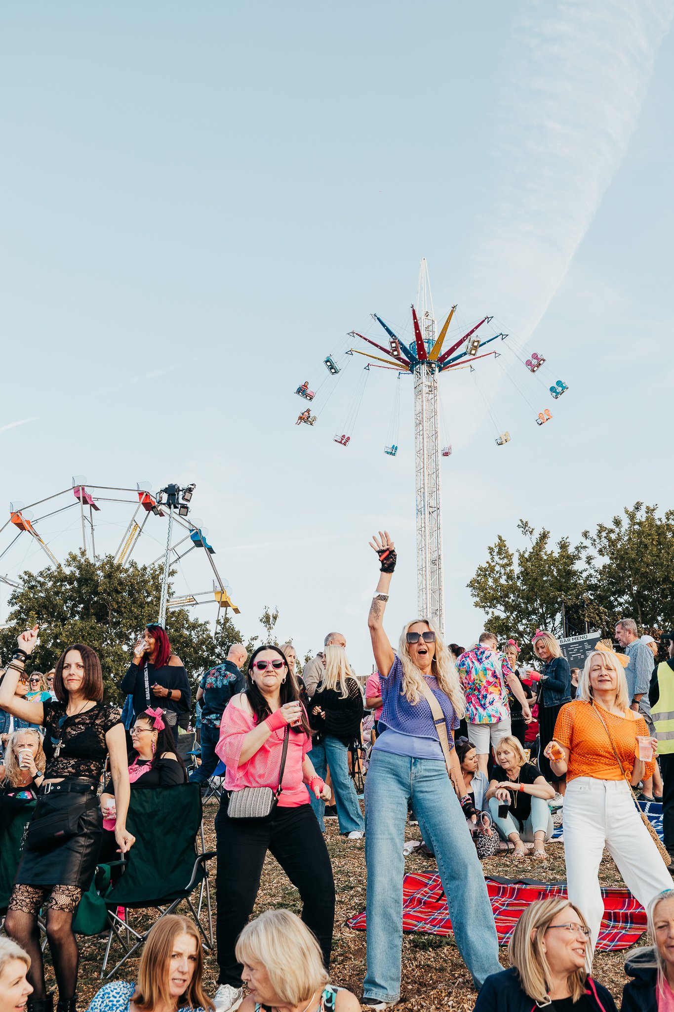 People enjoying a fairground with amusement rides, including swings and a colorful hanging ride, during daytime with a clear sky.
