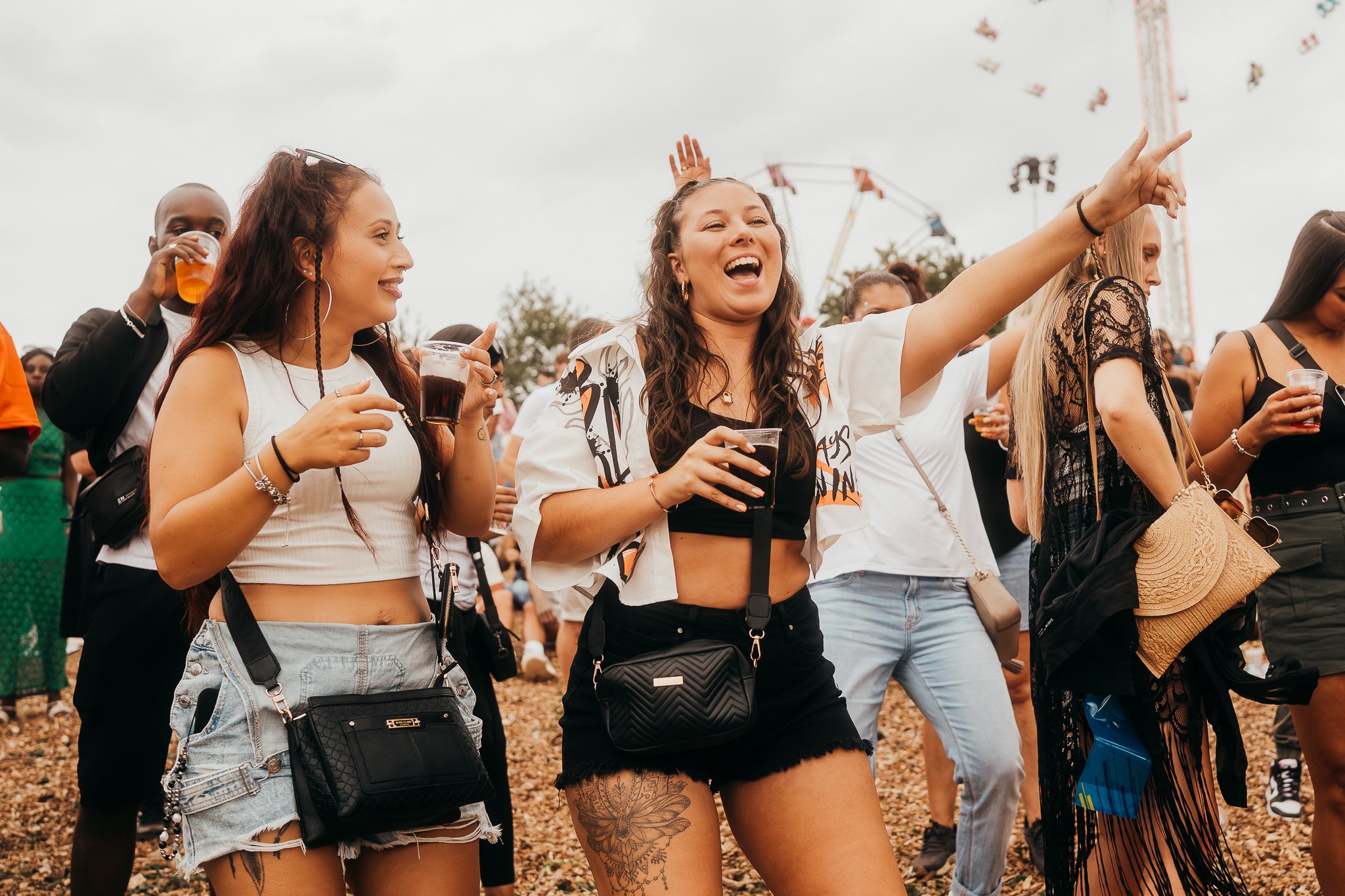 Group of people enjoying outdoor music festival, some drinking beverages, dancing, and smiling. Overcast sky, festival rides and decorations in background.
