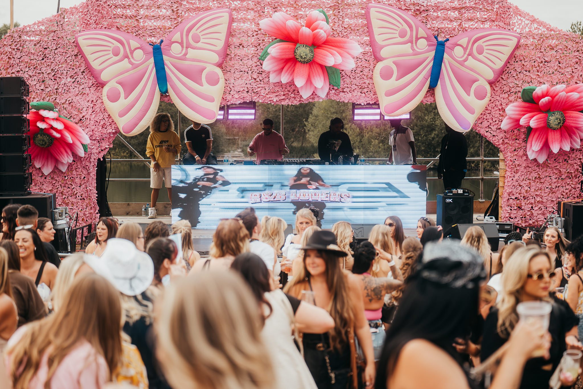 An outdoor music festival with a stage decorated with pink flowers and large butterfly decorations. A DJ is performing behind a digital screen, with a crowd of people dancing and enjoying the event.