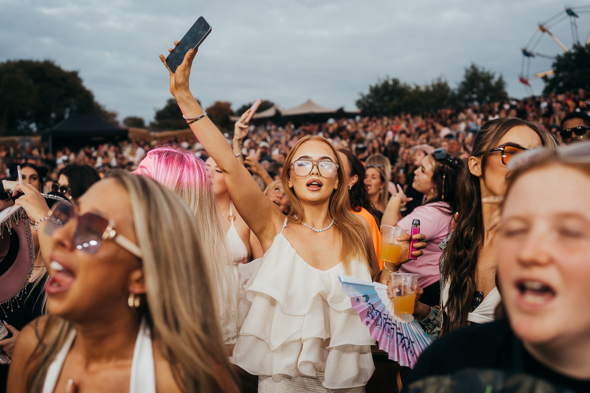 Crowd of people at an outdoor concert or festival, with many holding drinks and taking photos, some wearing sunglasses and summer clothing.