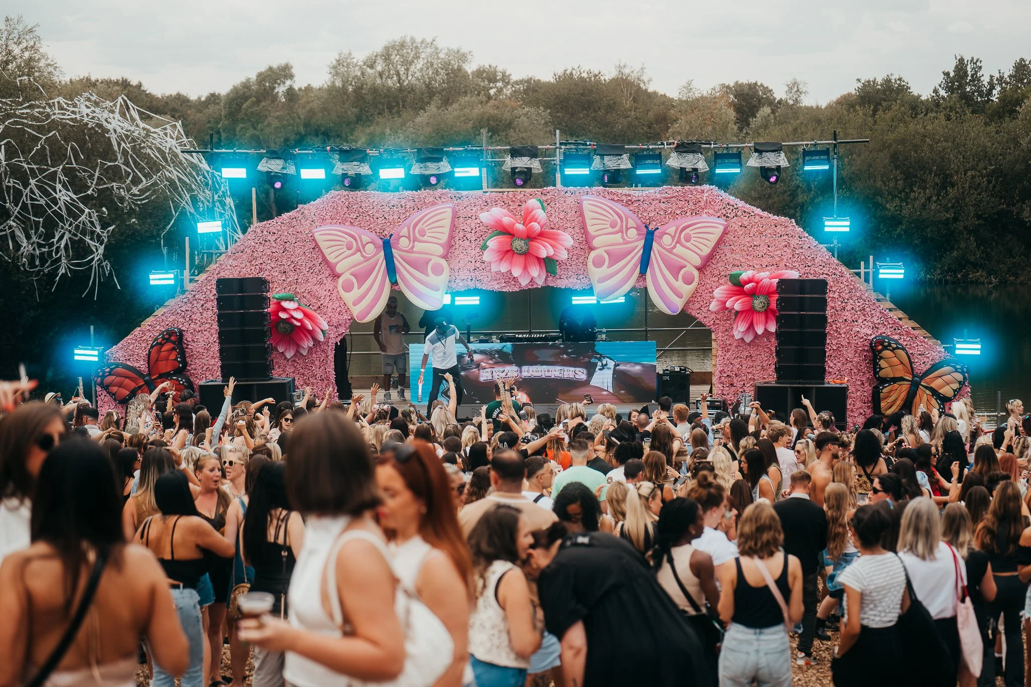 Large outdoor party or music festival near a body of water with a decorated stage featuring pink flowers and butterfly ornaments, crowd of people dancing and socializing in front of the stage.