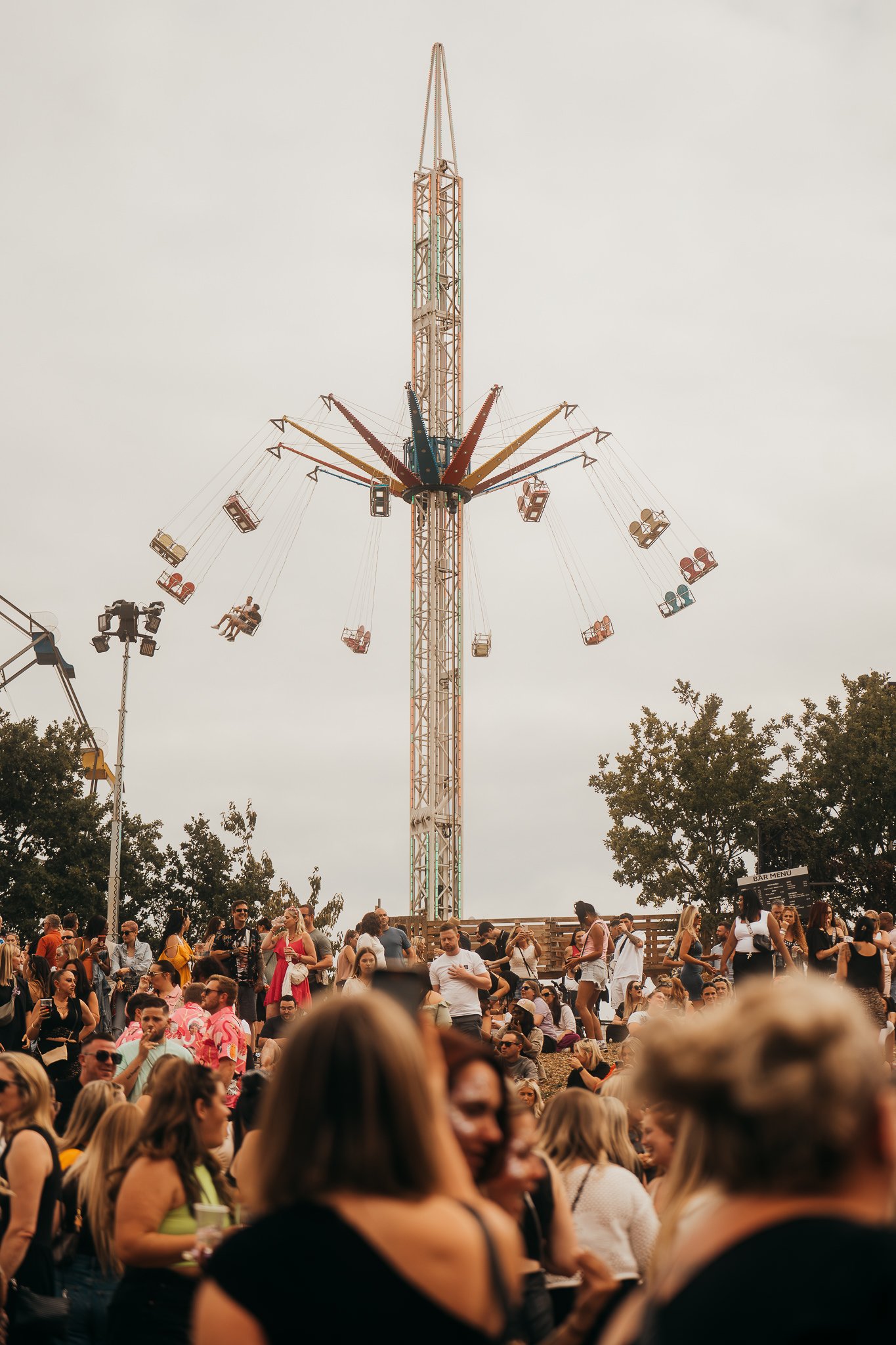 A large crowd of people at an outdoor fair or carnival with a swing ride in the background, where swings are suspended from a tall tower.