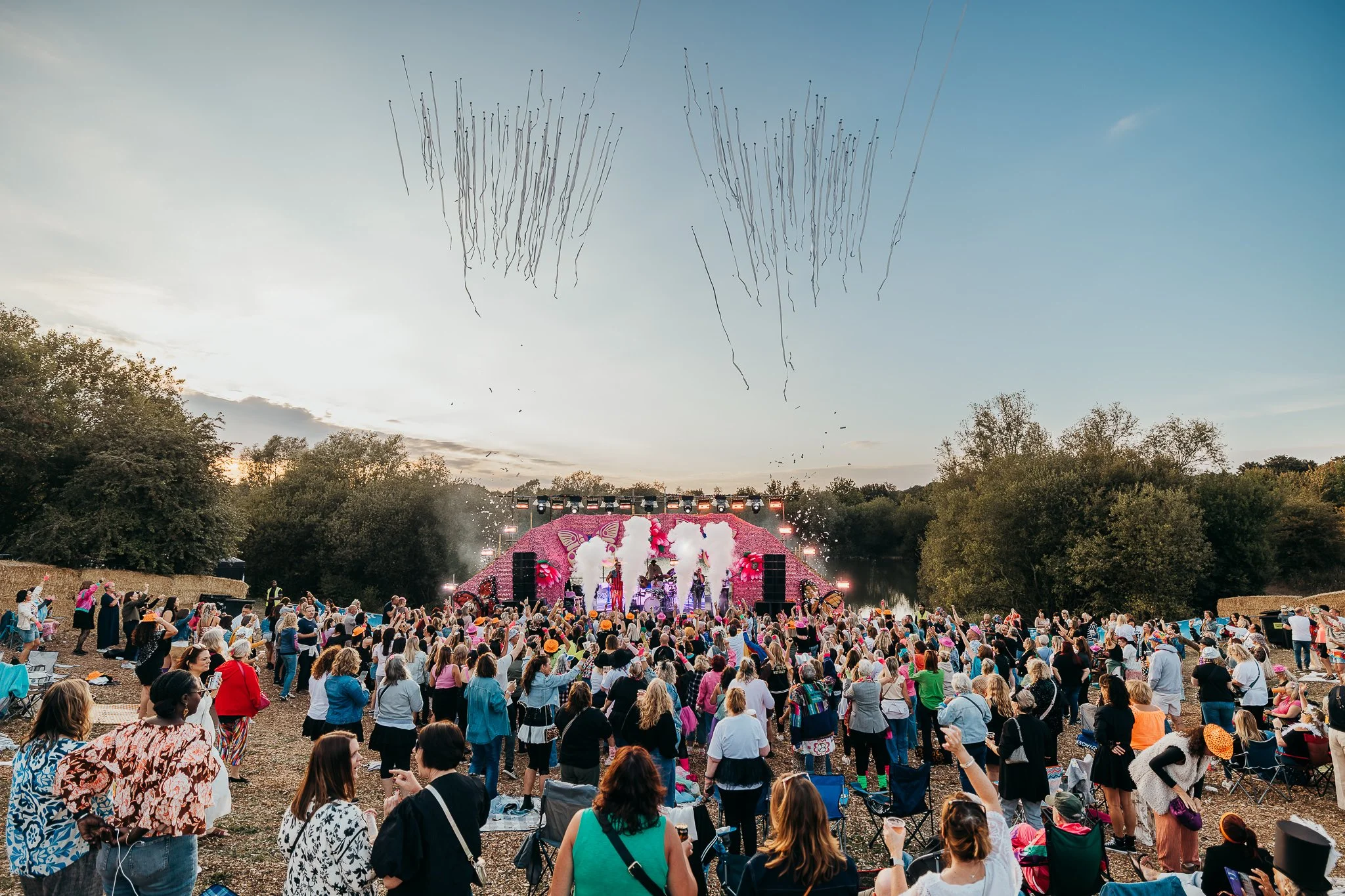 Large outdoor concert with a crowd gathered in front of a colorful stage decorated with pink flowers and butterfly designs, set near a river with trees in the background during sunset.