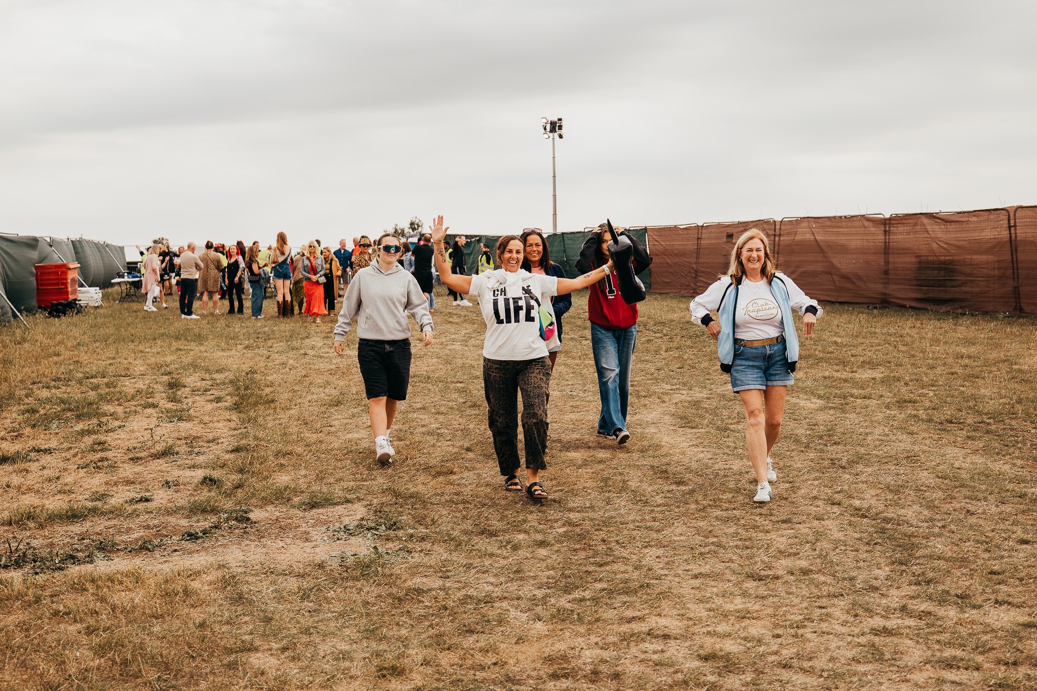 Group of women walking in a field at an outdoor event with tents and a cloudy sky in the background.