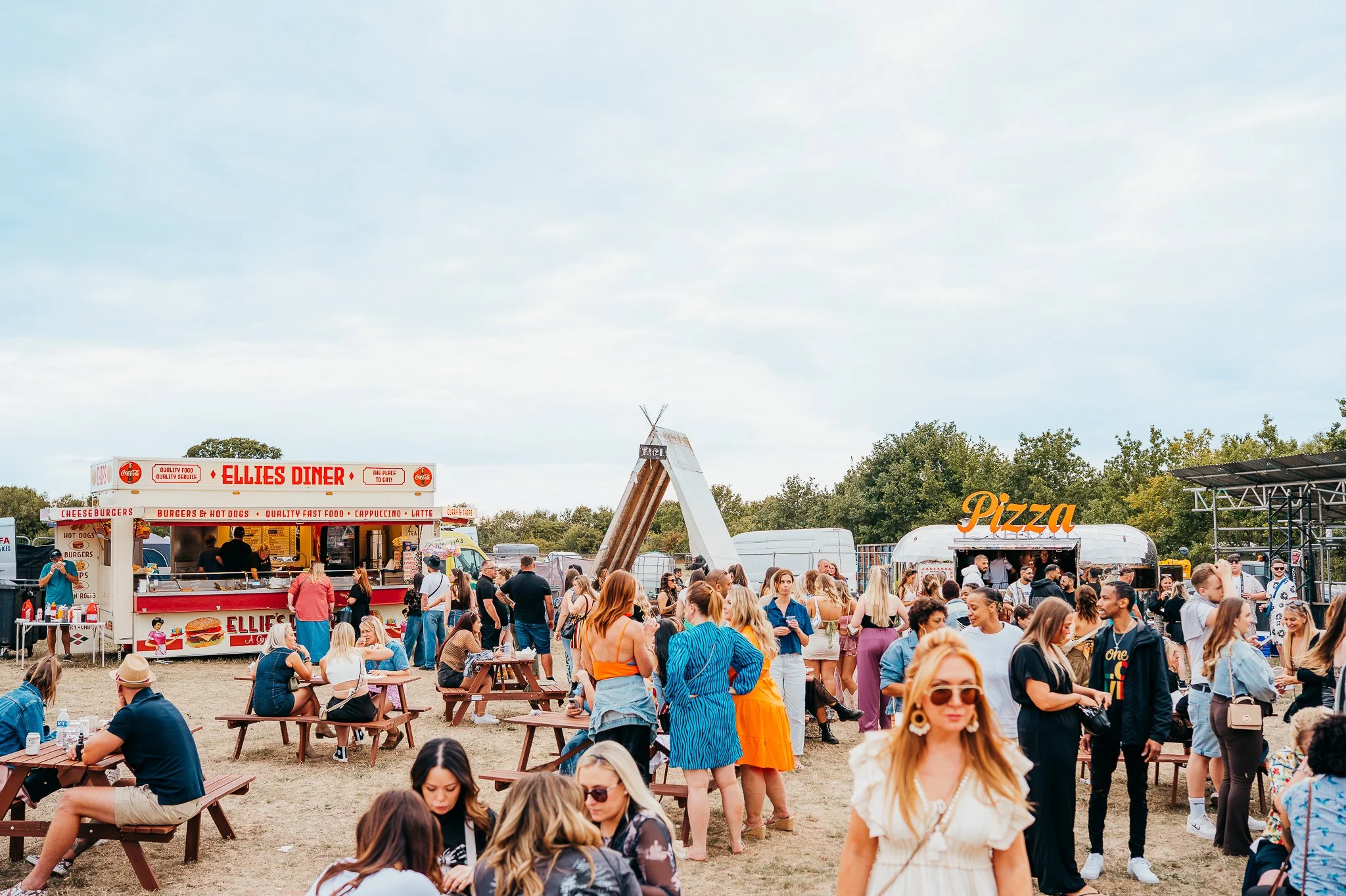 People at an outdoor food festival with food trucks labeled "Ellie's Diner" and "Pizza" and a large wooden teepee structure in the background.