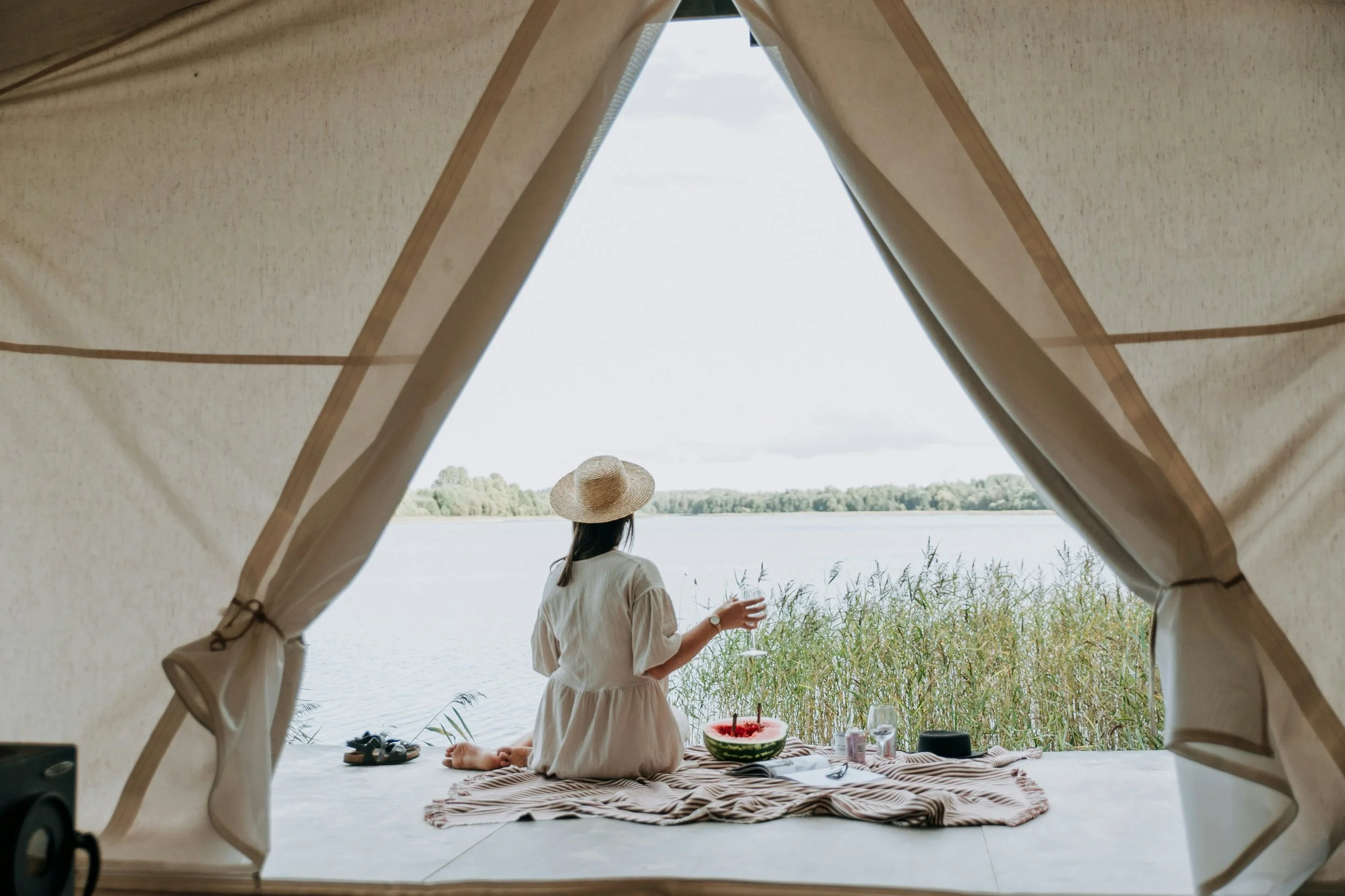 A woman sitting on a blanket by a lake, wearing a wide-brimmed hat, and holding a glass of wine, with a watermelon-shaped cake and other items on the blanket, viewed from inside a tent.