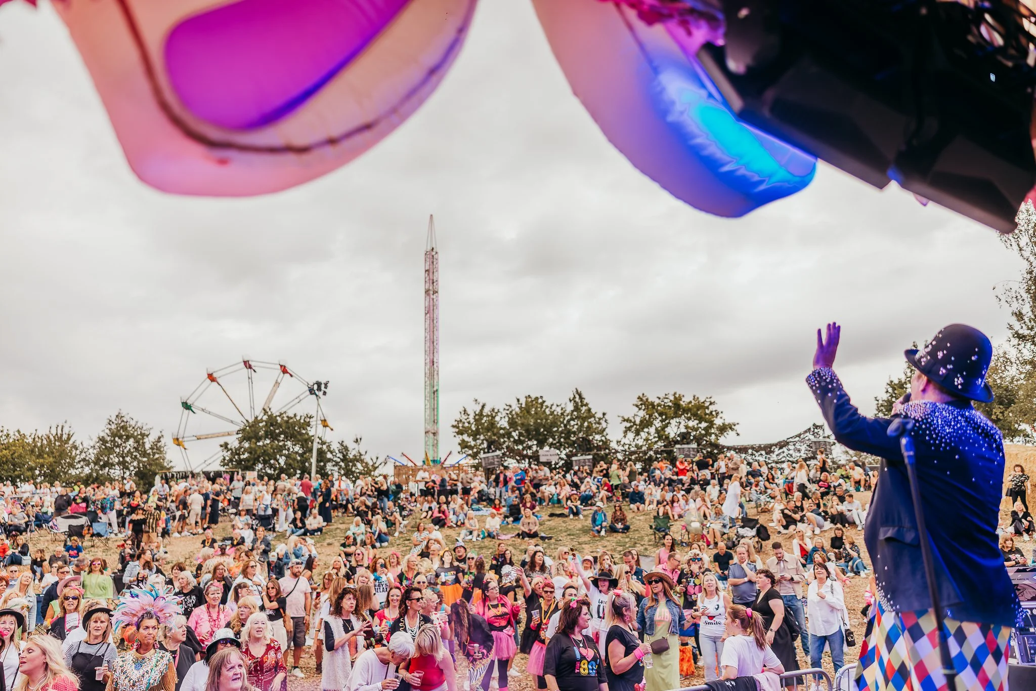 Crowd gathered at an outdoor event under cloudy sky, with a performer dressed as a clown or mime waving to the audience, carnival rides in the background, and colorful balloons or decorations overhead.
