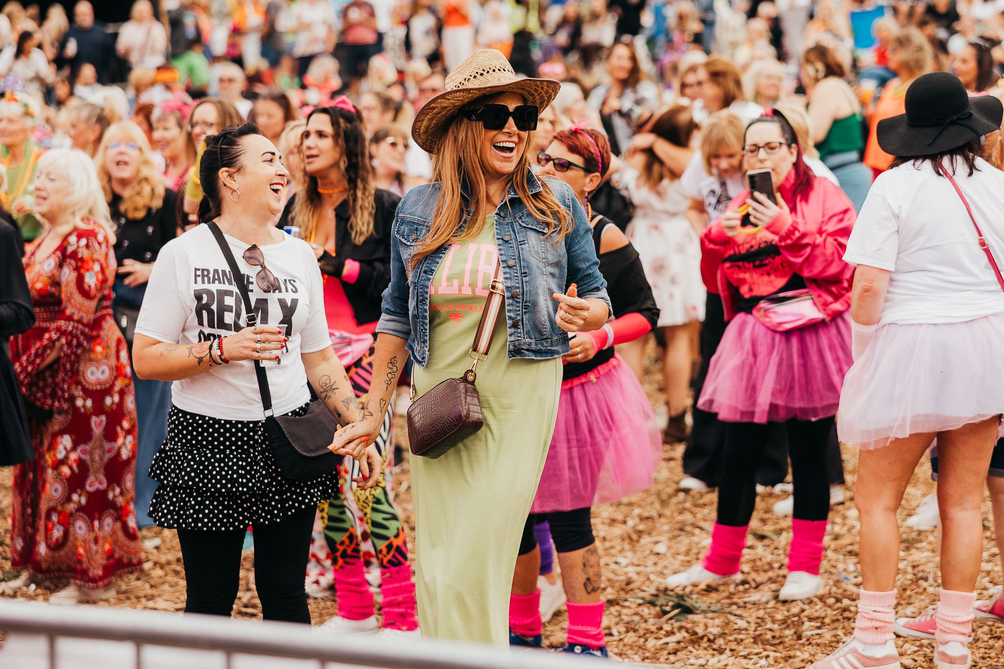 A group of people at a crowded outdoor event, many wearing colorful pink tutus and casual clothing, some taking photos or holding hands, with chalk on the ground and a mix of smiling, laughing, and chatting individuals.