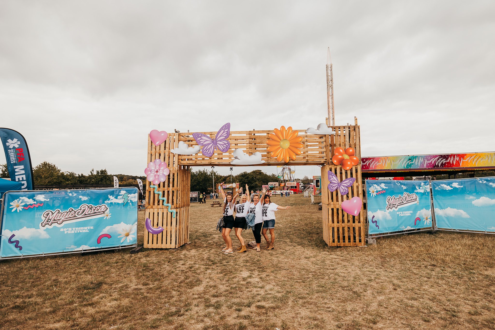 Four women wearing white t-shirts with 'IF' standing under a decorated entrance at an outdoor fair with festival booths and rides in the background