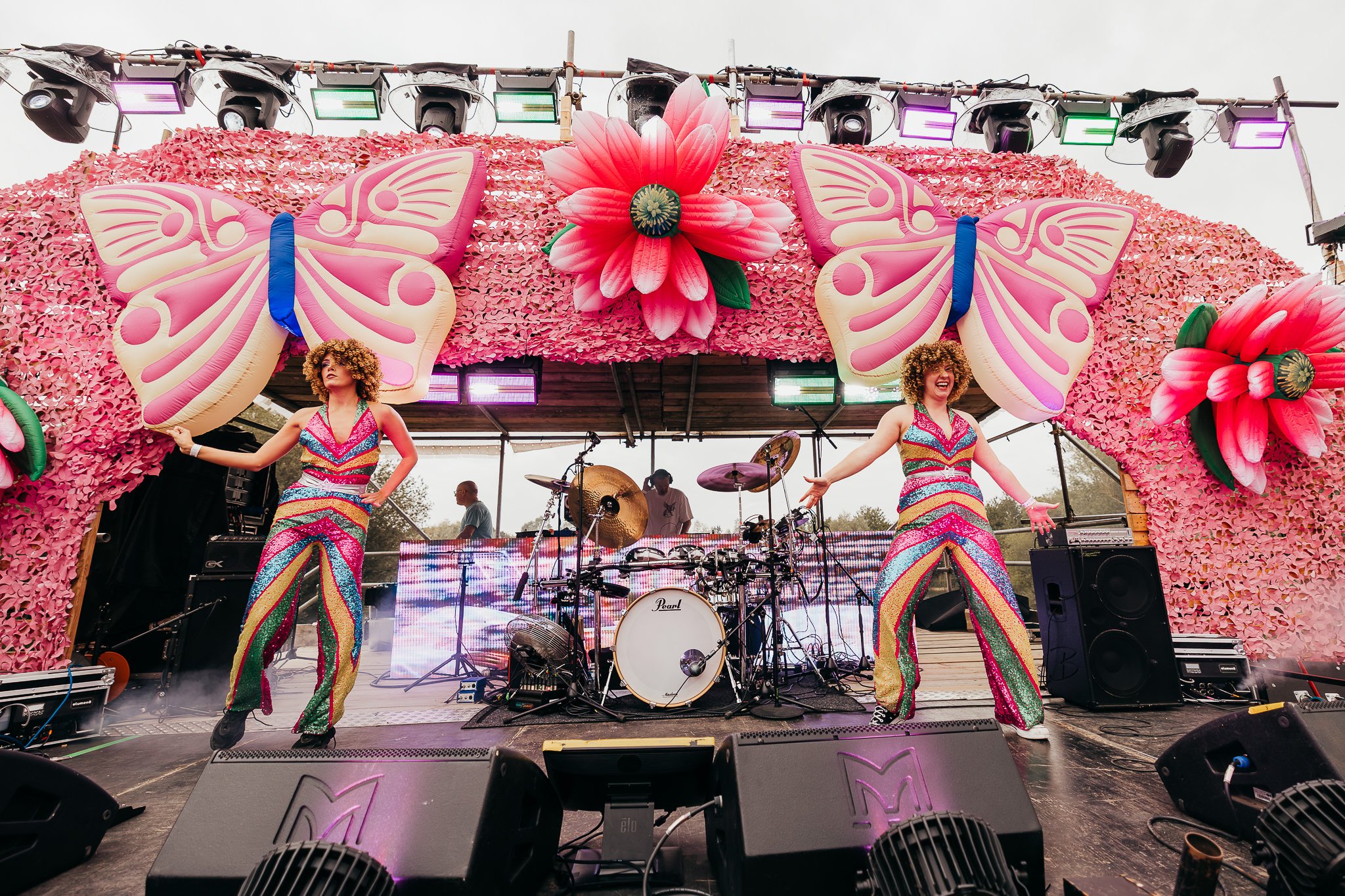 Two performers in colorful, striped jumpsuits dancing on a stage decorated with large pink daisies and butterfly decorations, with a drum set and dj behind them, during an outdoor event.