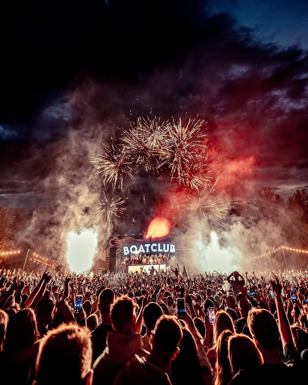 Crowd at an outdoor music festival with fireworks in the sky, a stage with a sign that says 'BOATCLUB', and many people holding phones up to record the event.