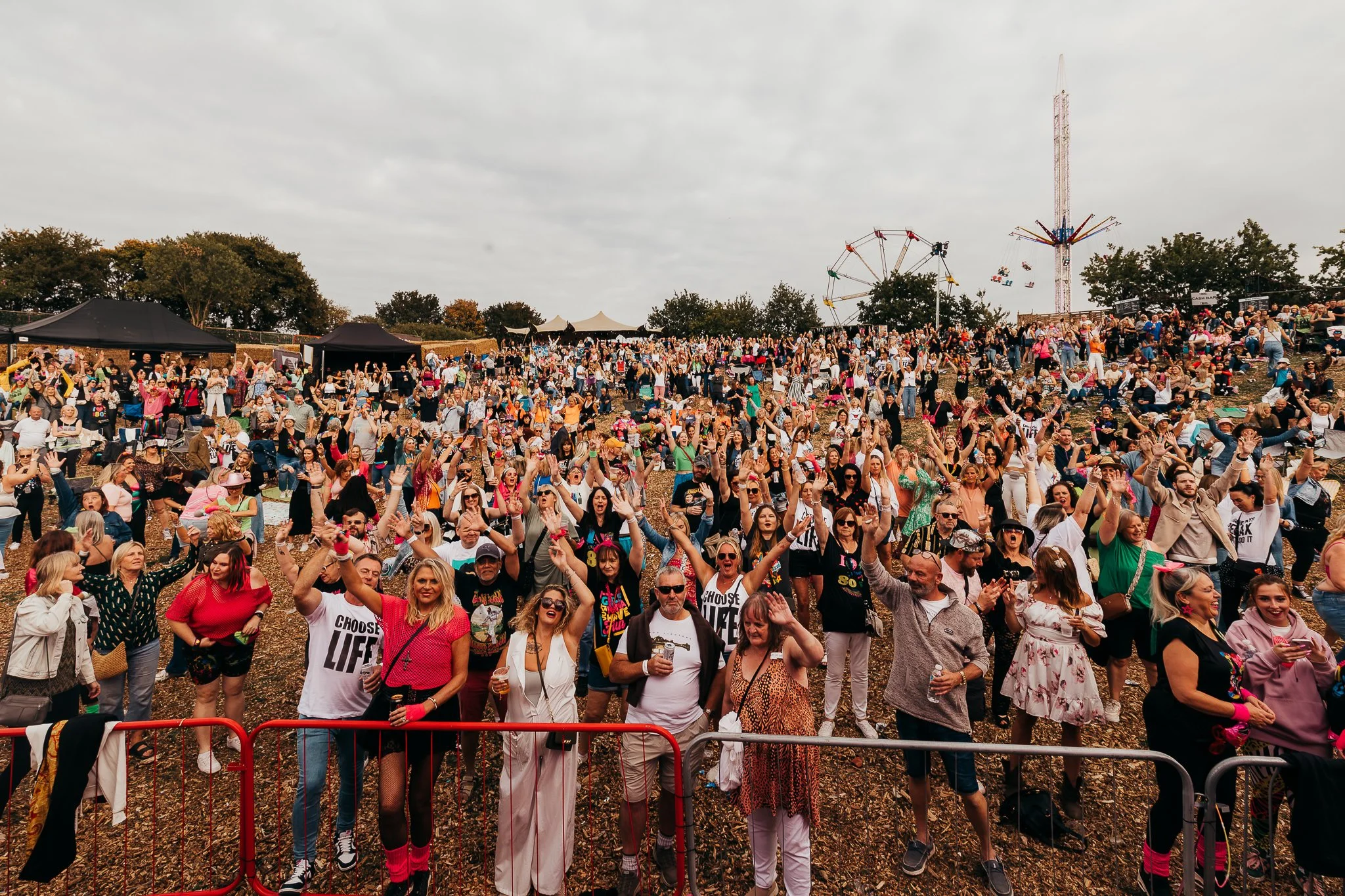 Crowd of people at outdoor festival with amusement rides and tents in background.