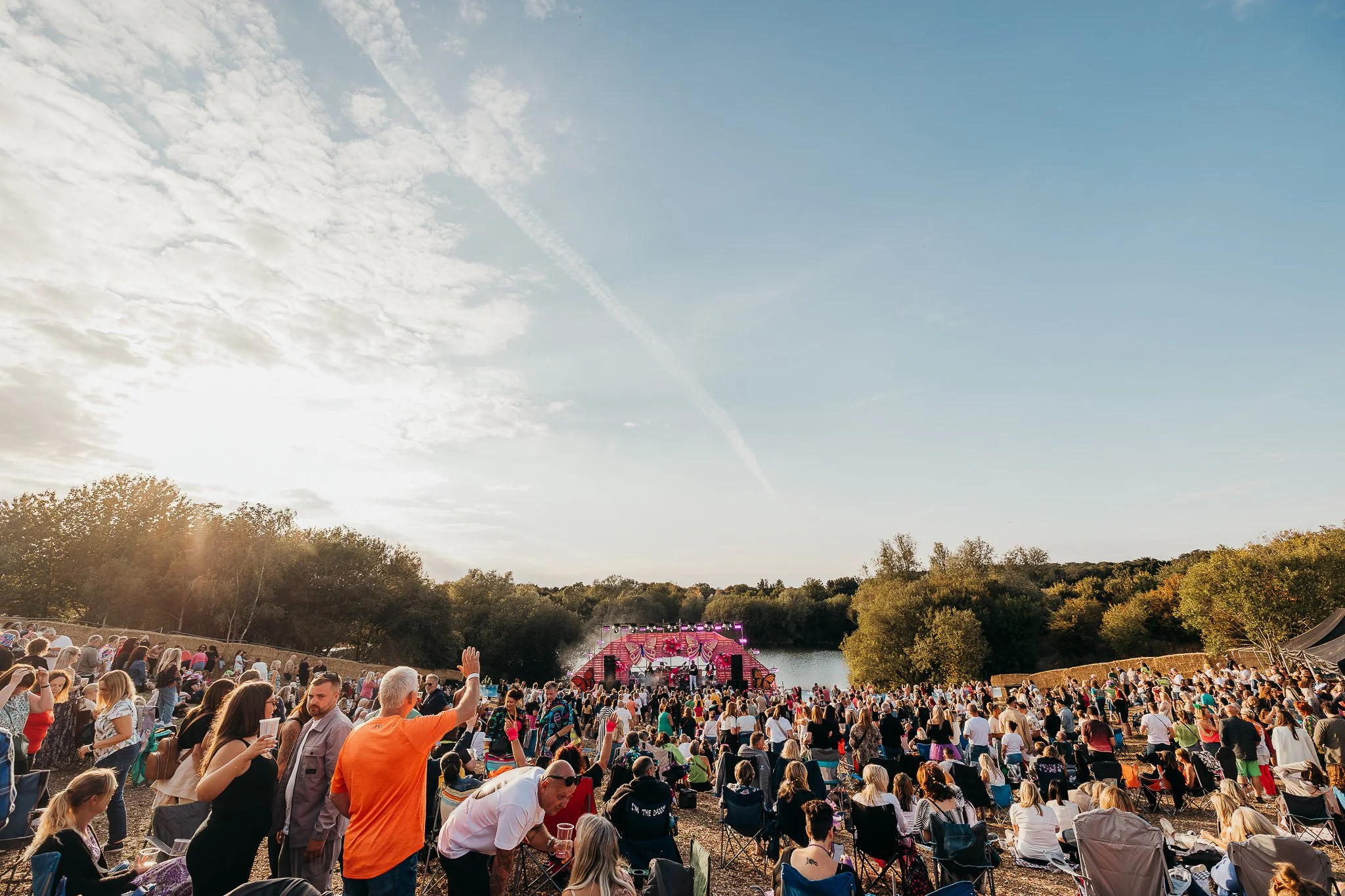 Outdoor music festival with a large crowd by a lake, stage with pink decorations, trees surrounding the area, during sunset.