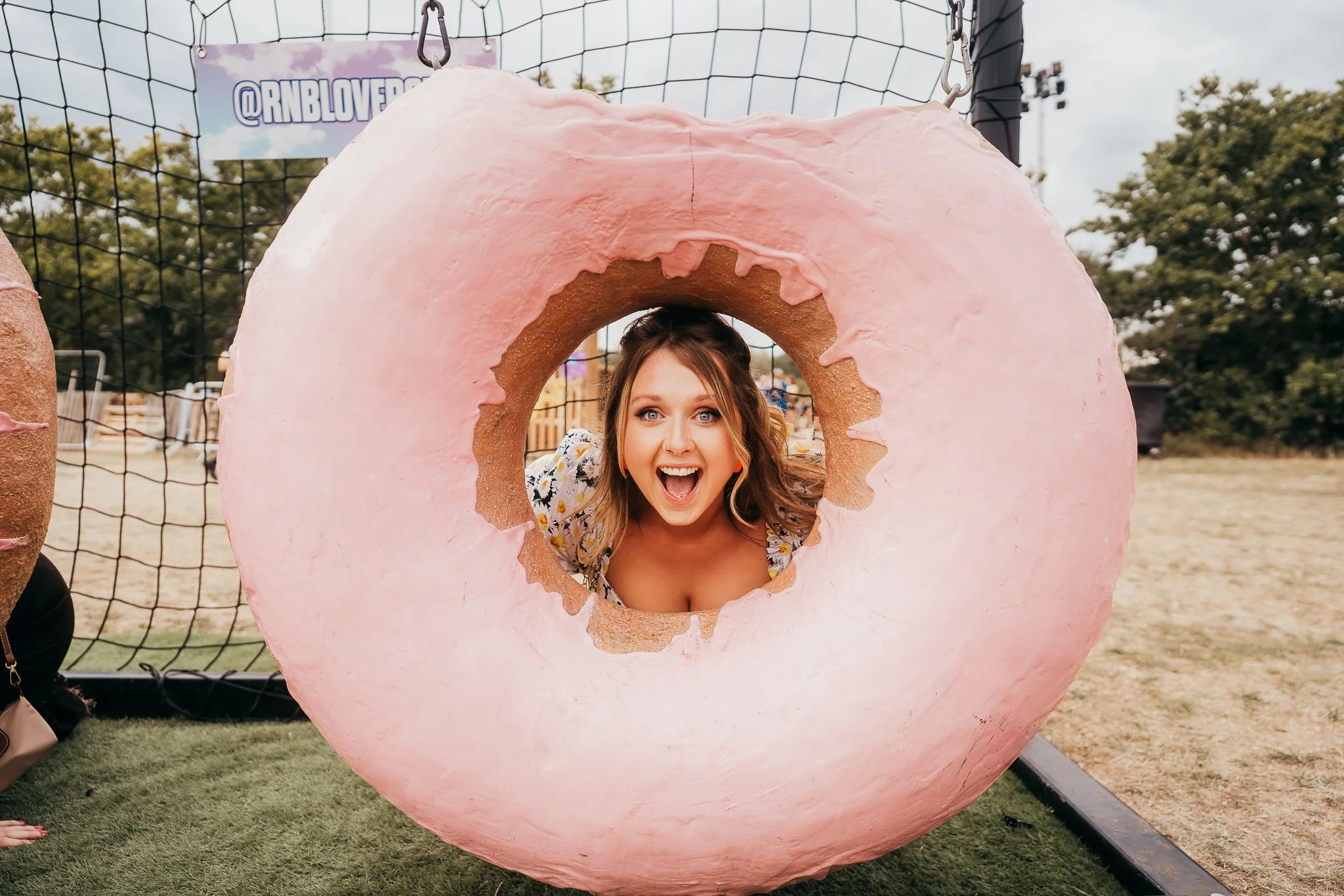 A woman smiling and peeking through the center hole of a giant, pink donut-shaped sculpture with light pink icing and brown base at an outdoor event or park.