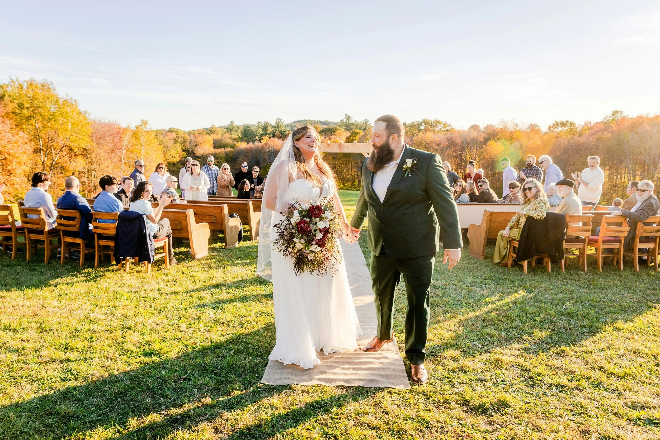 A bride and groom holding hands and smiling at each other during an outdoor wedding ceremony in a scenic setting with trees displaying fall colors. Guests are seated on wooden benches and standing in the background, and the sunlight creates a warm, joyful atmosphere.