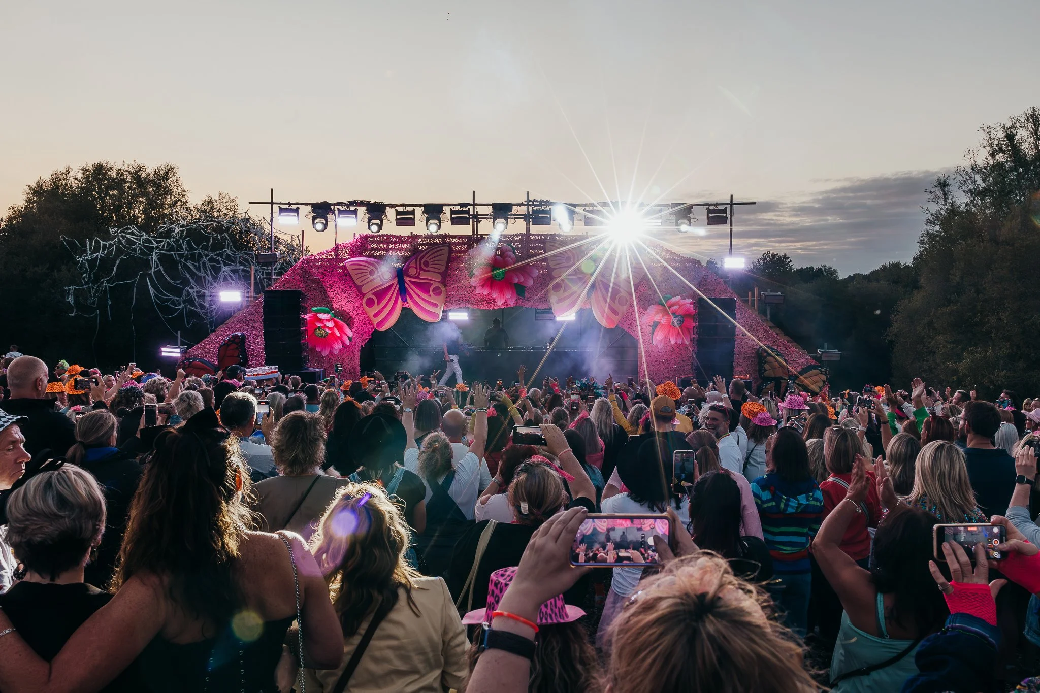 Crowd of people attending an outdoor concert or festival at sunset, with a decorated stage featuring large butterfly and flower decorations, and bright stage lights.