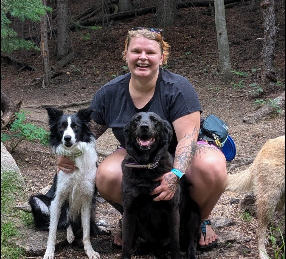A woman smiling outdoors, kneeling with two dogs on a forest trail.