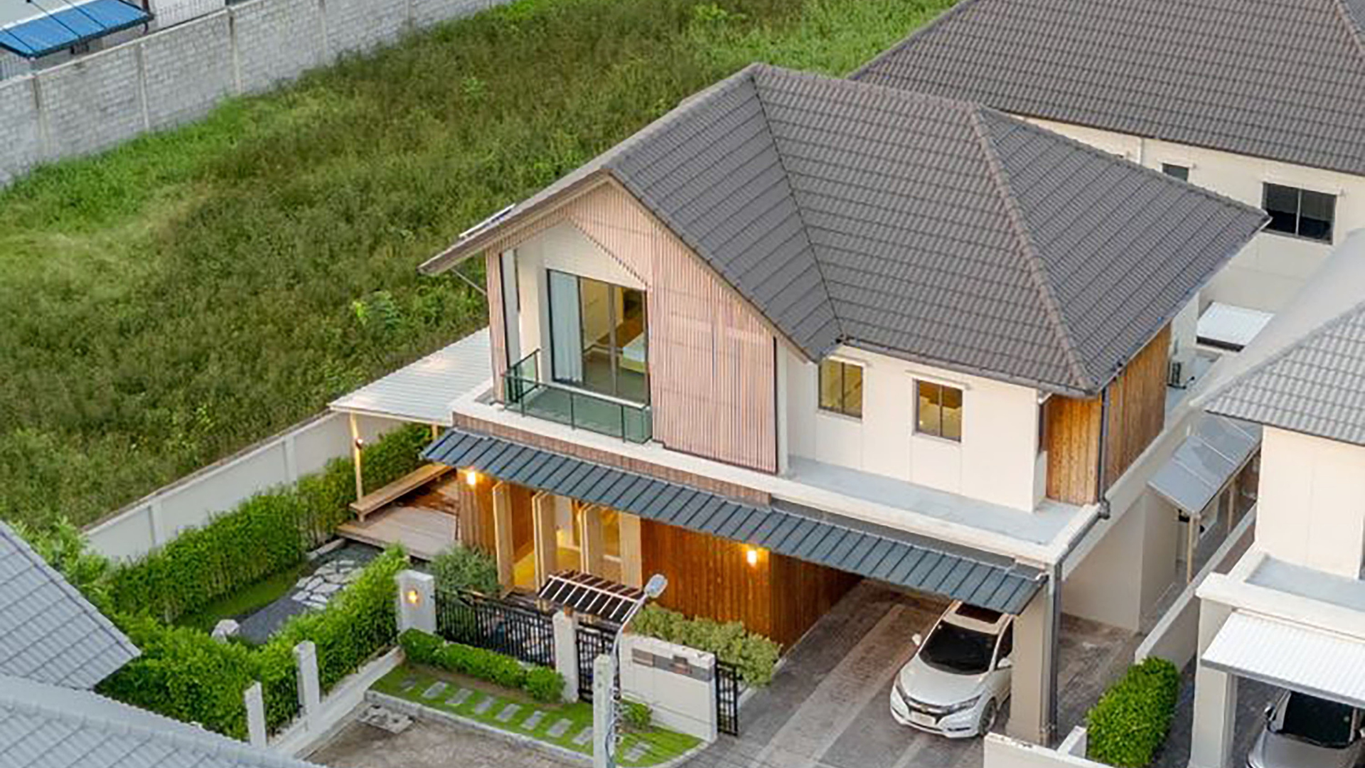 A modern two-story house with a tiled roof, wooden and white exterior walls, a small balcony, a driveway with a white car, and a backyard with greenery and outdoor lighting.