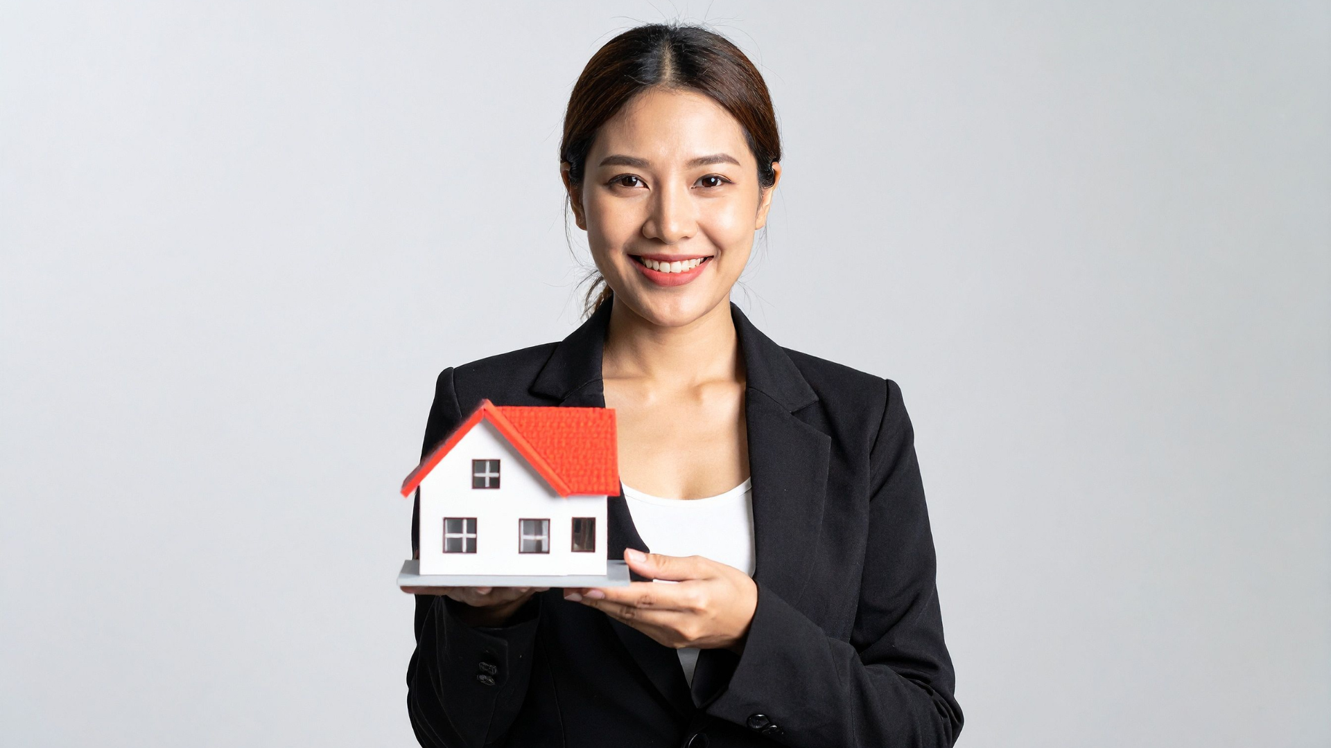 A woman in a black blazer holding a small model house with a red roof and white walls, smiling at the camera.