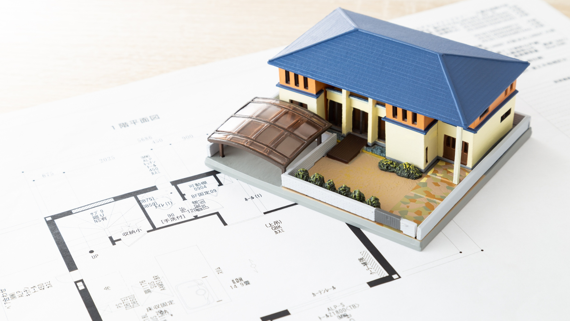 A detailed miniature model of a house with a blue roof, a carport with a transparent roof, and a small landscaped yard, placed on top of architectural blueprints.