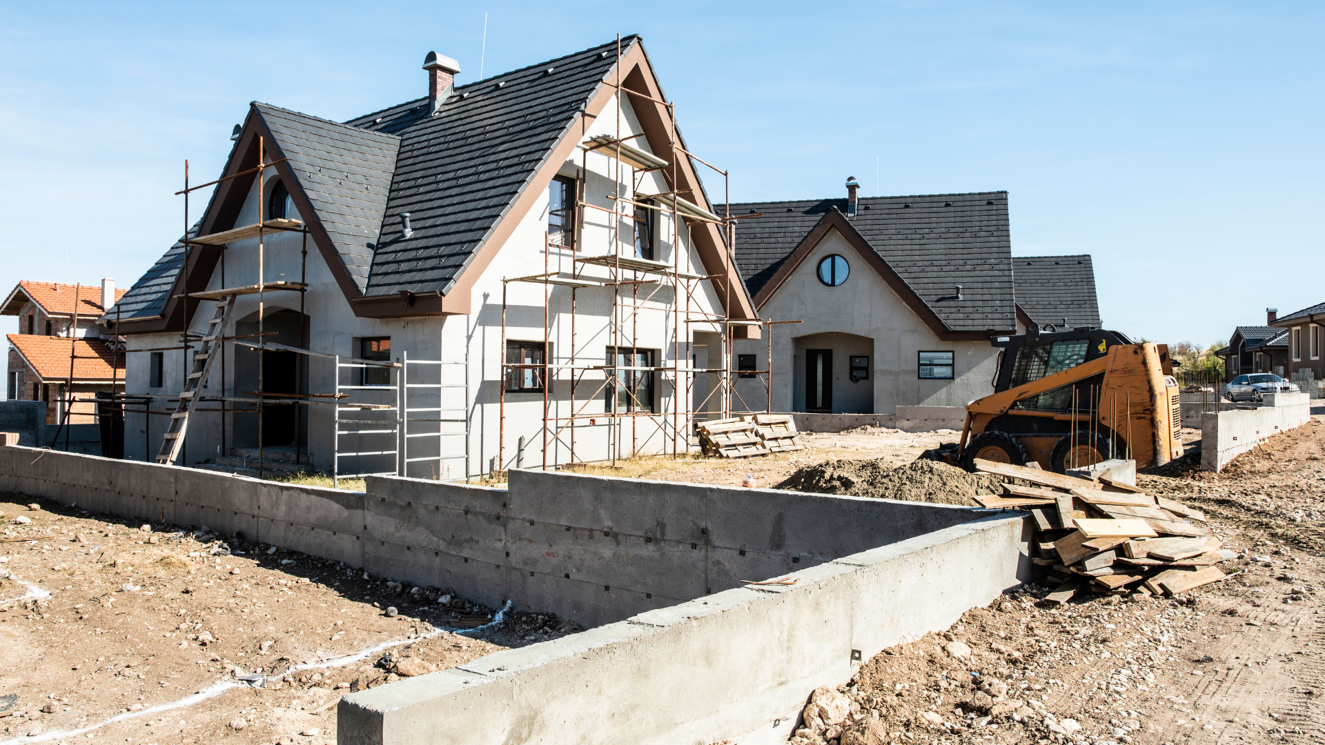 A house under construction with scaffolding, a small retaining wall, and construction equipment on a dirt lot.