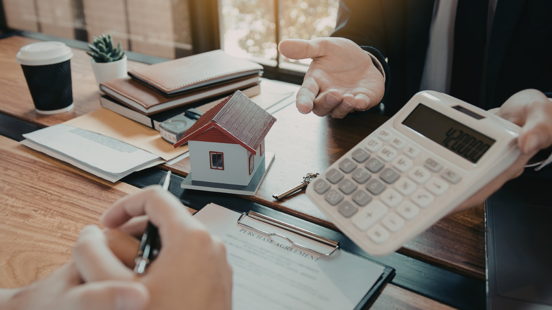 A person is signing a purchase agreement at a table with a small house model, a calculator showing 420000, and office supplies, during a real estate transaction.