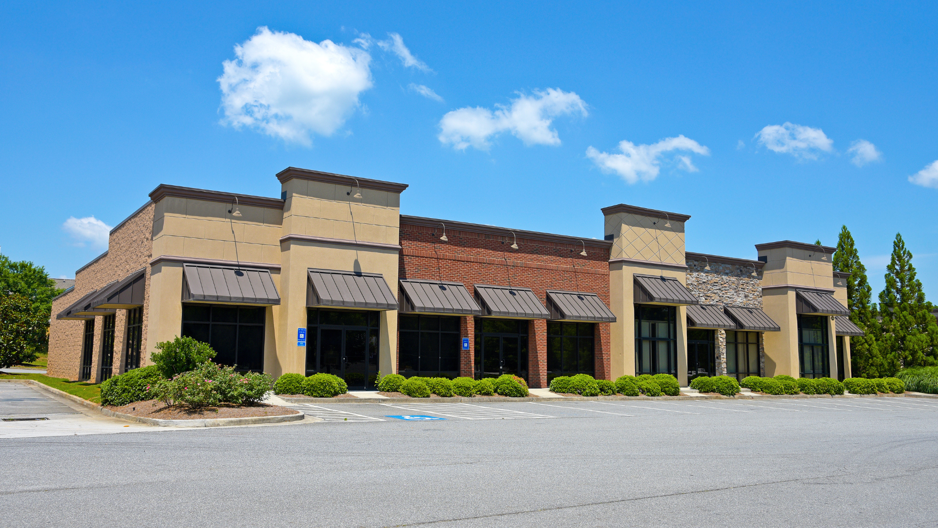 Empty commercial building with beige and brick facades, black windows, multiple entrances with black awnings, surrounded by landscaping and a parking lot under a blue sky with clouds.