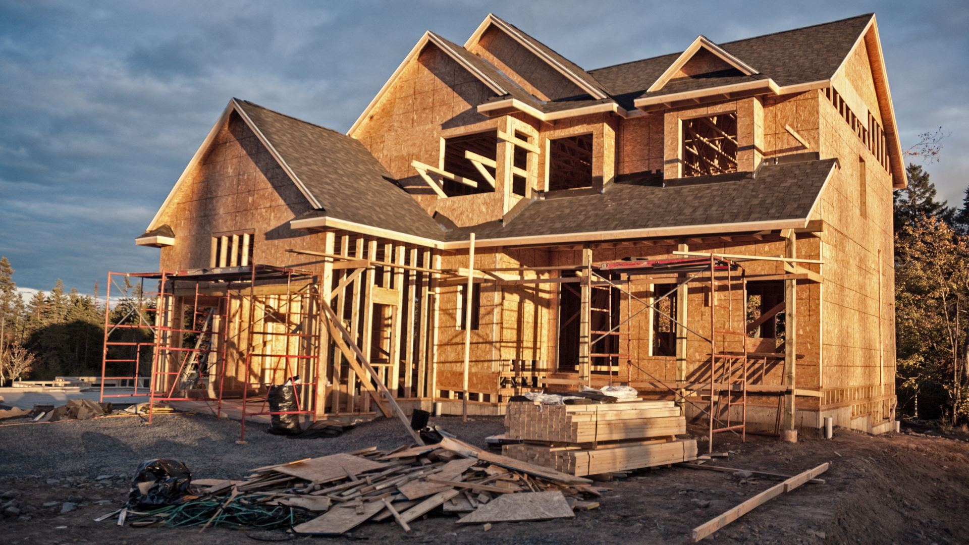Under construction two-story house with wooden framing and scaffolding, existing roof shingles, and construction materials in front.