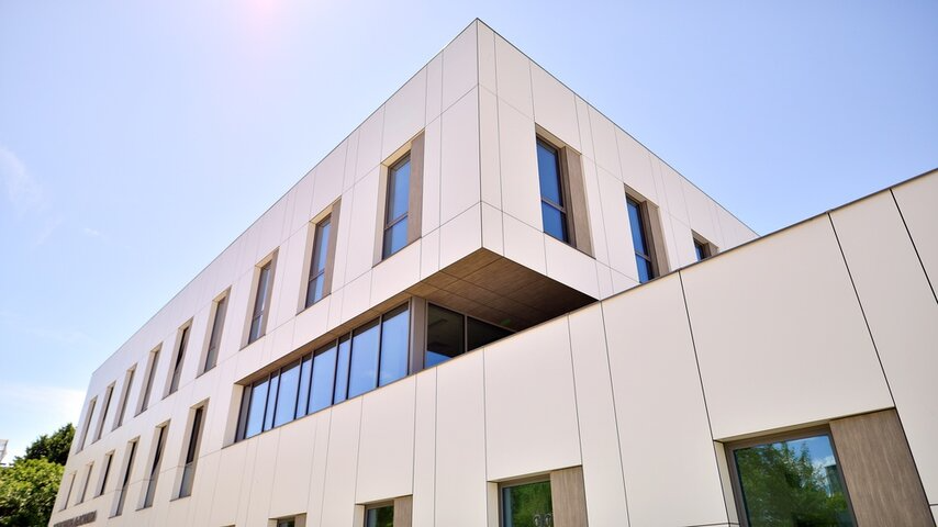 A modern, white multi-story building with large windows and a flat roof, taken from a low angle against a clear blue sky.