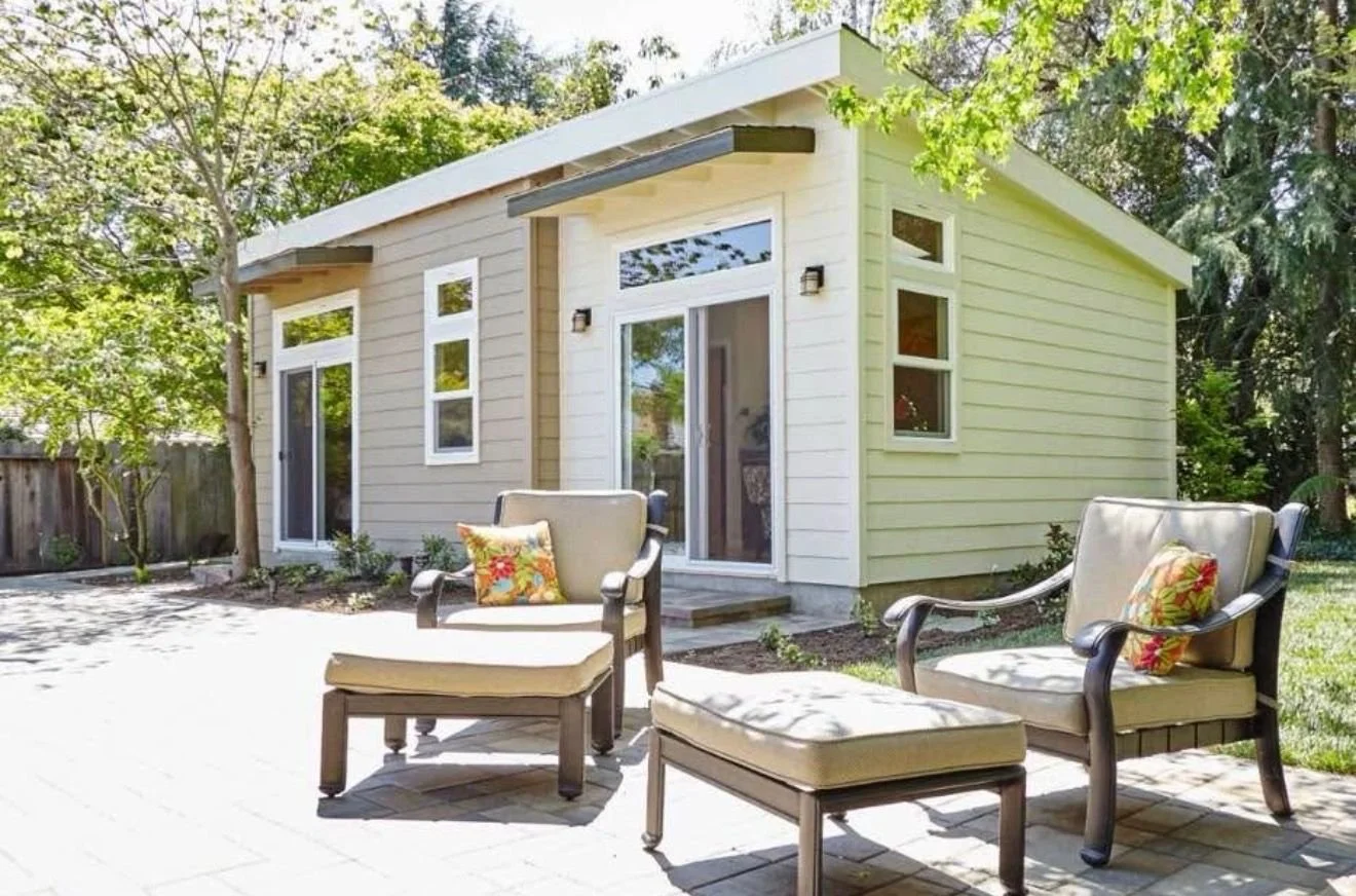 A backyard patio with two cushioned chairs and an ottoman, in front of a small beige house with white trim, surrounded by green trees and shrubbery.