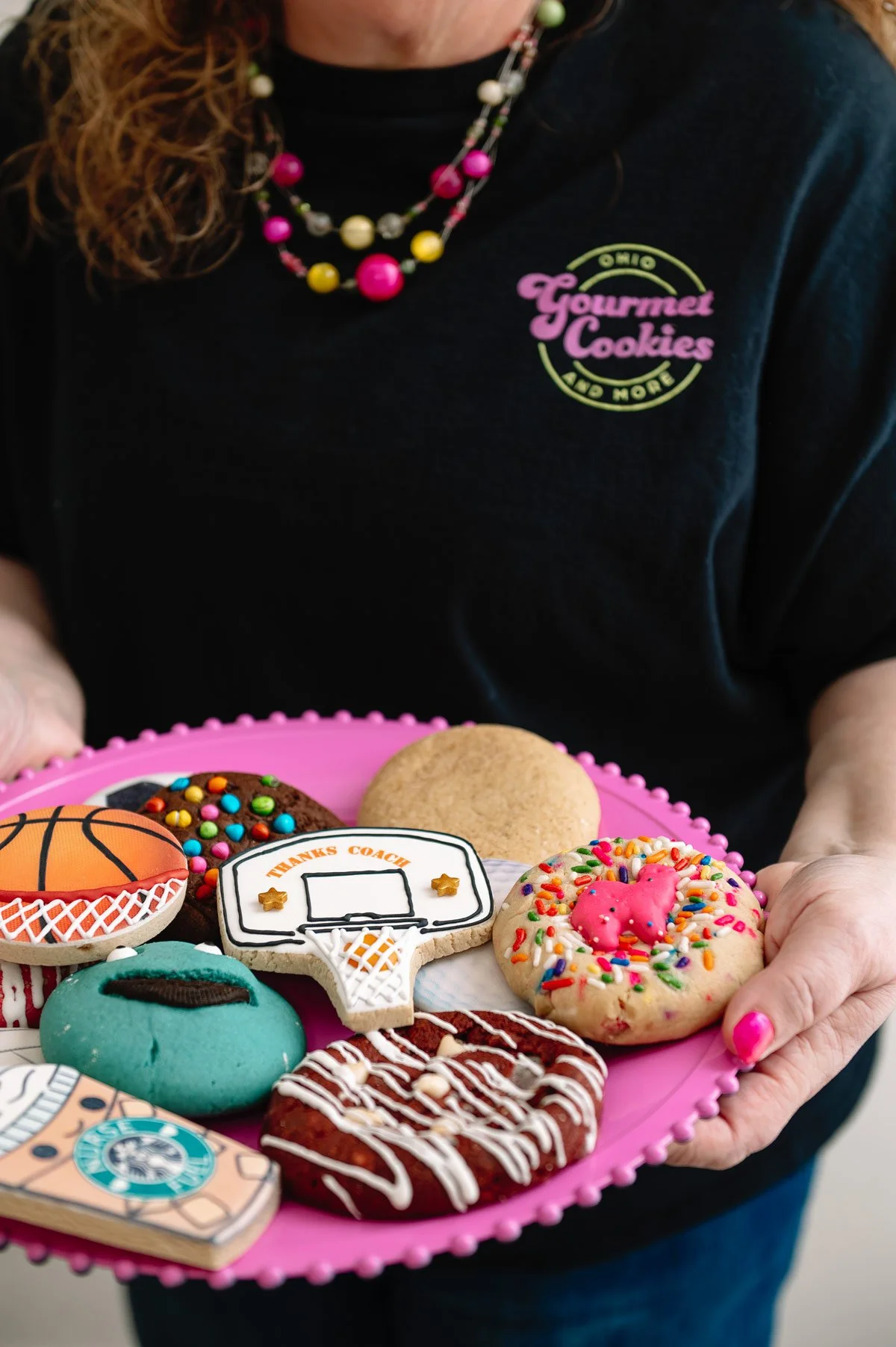 Person wearing a black shirt and colorful beaded necklace holding a pink tray with decorated cookies, including a basketball, a cupcake, a football, a donut, a cookie with a basketball hoop saying 'Thanks Coach,' and a cookie with a pink pig on top.