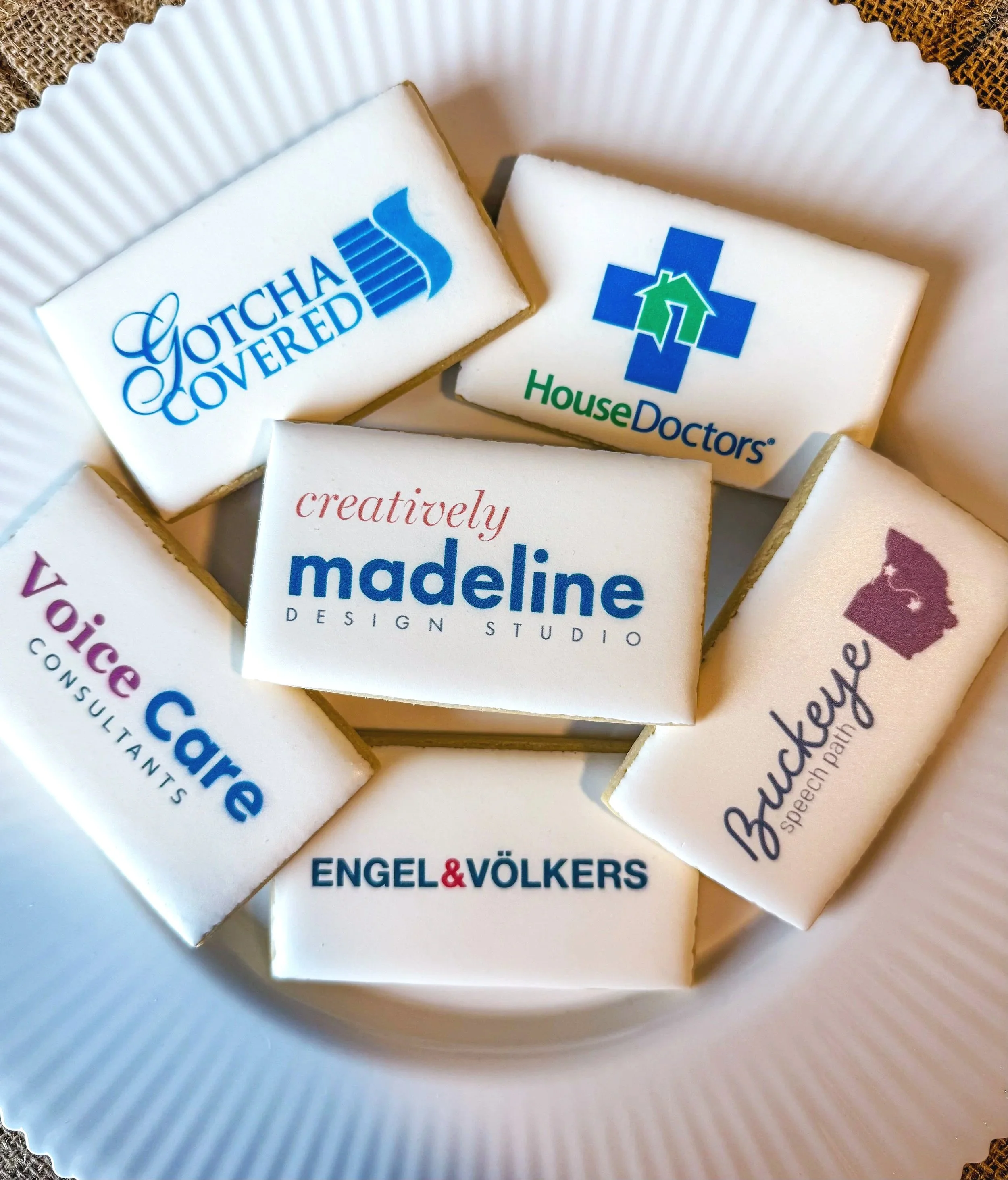 Collection of cookies with various company logos and branding, including Gotcha Covered, House Doctors, Creatively Madeline, Voice Care Consultants, Buckeye Speech Path, and Engel & Völkers, arranged on a white paper plate.