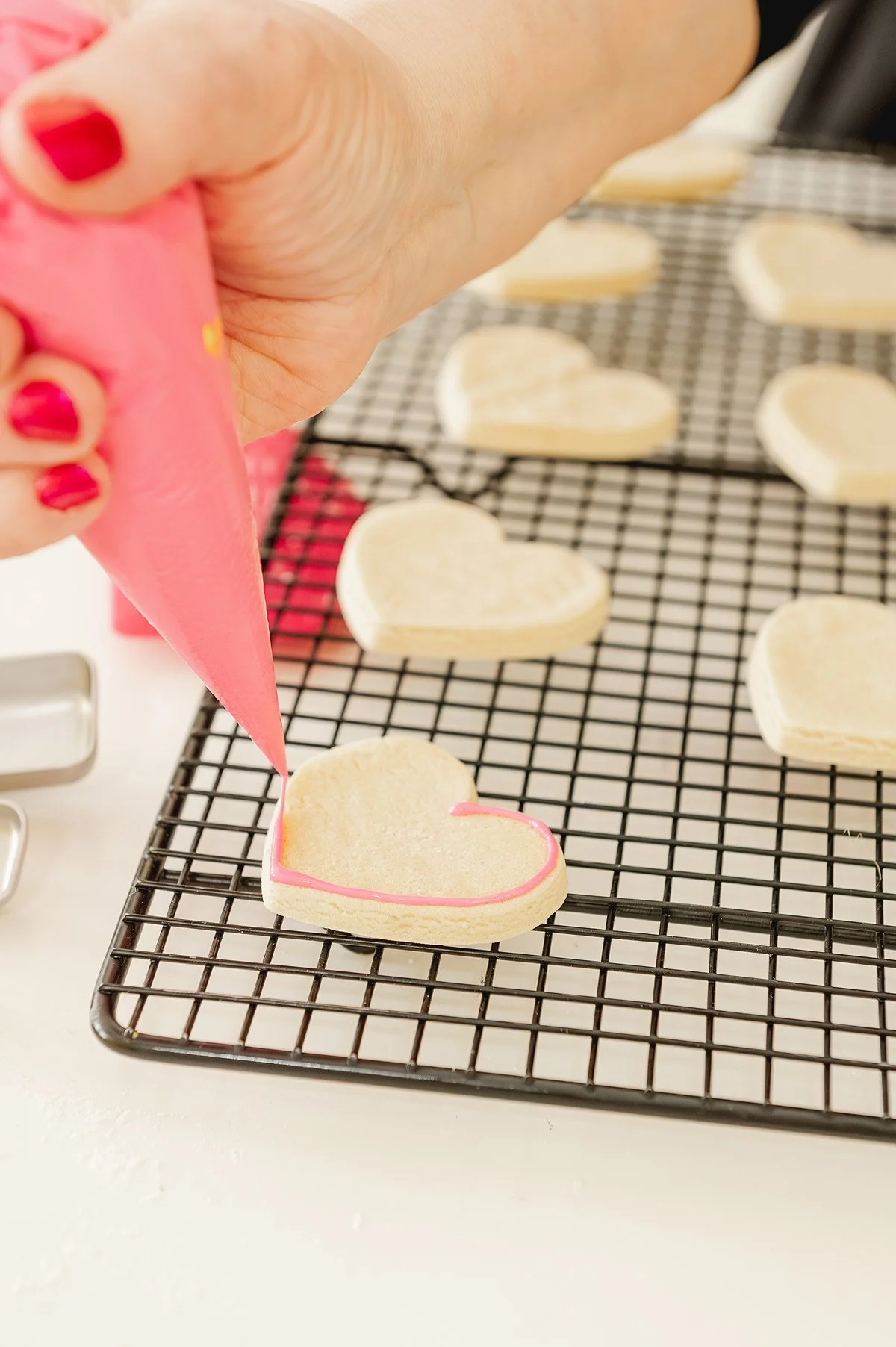 Person piping pink icing onto a heart-shaped cookie on a cooling rack.