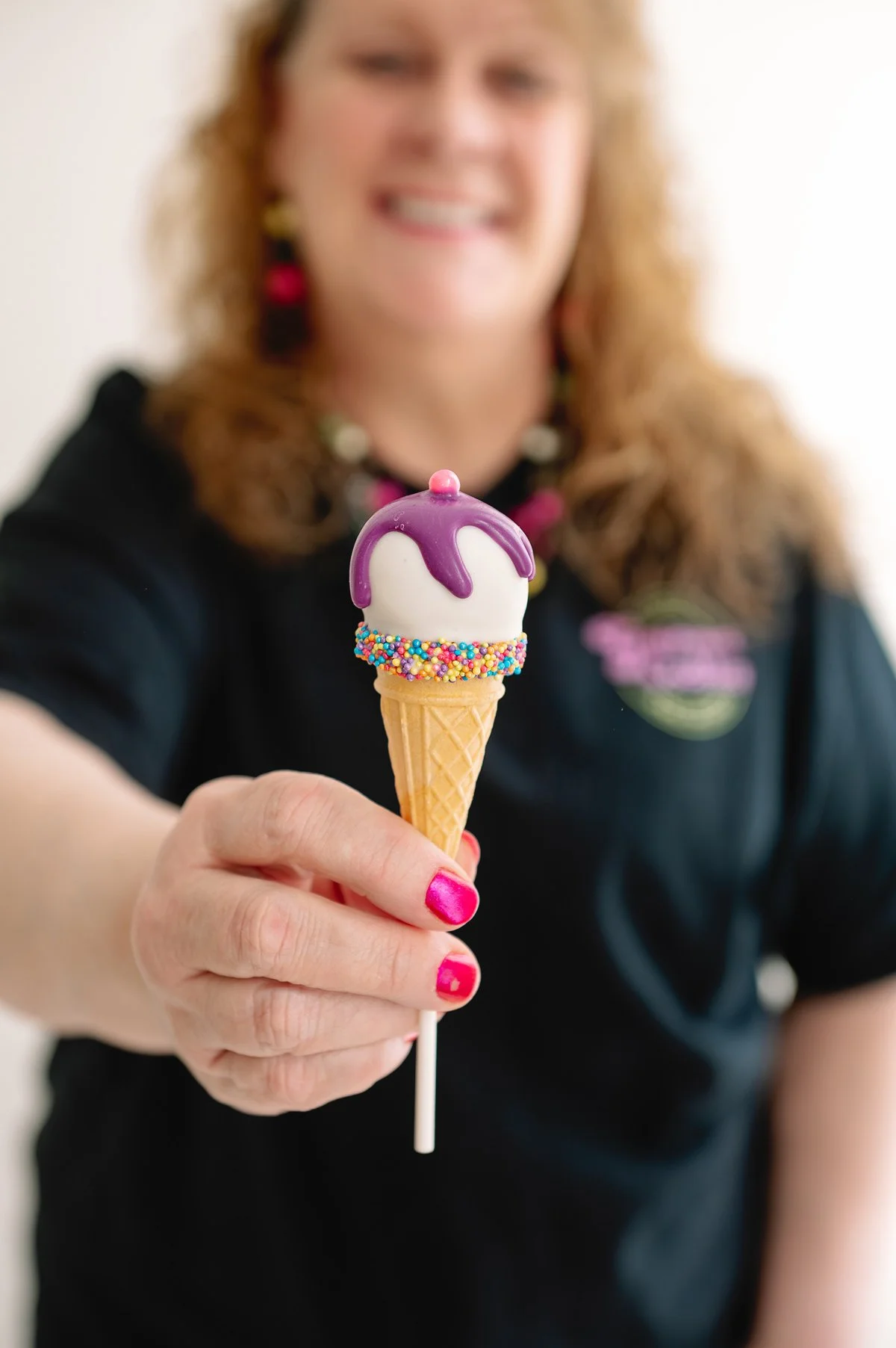 A woman holding a small ice cream cone with purple and white ice cream topped with purple syrup and colorful sprinkles, smiling in the background.