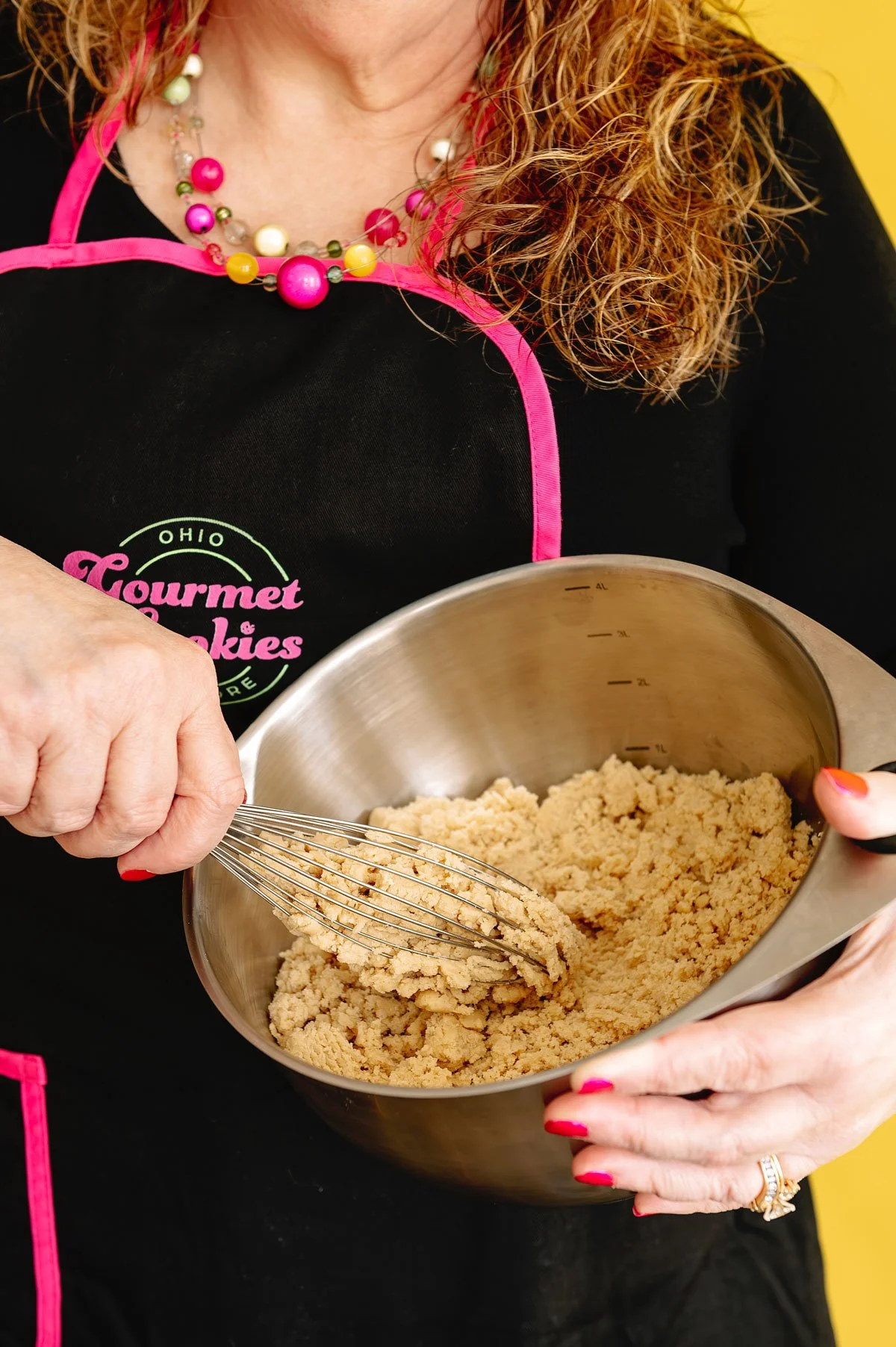 A woman with curly red hair wearing a black apron with pink trim and a colorful beaded necklace, whisking cookie dough in a metallic mixing bowl.